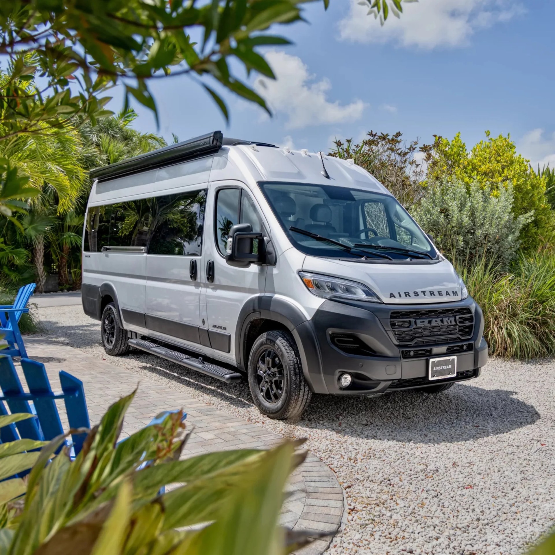 Silver Airstream camper van with black trim parked on gravel near tropical plants.