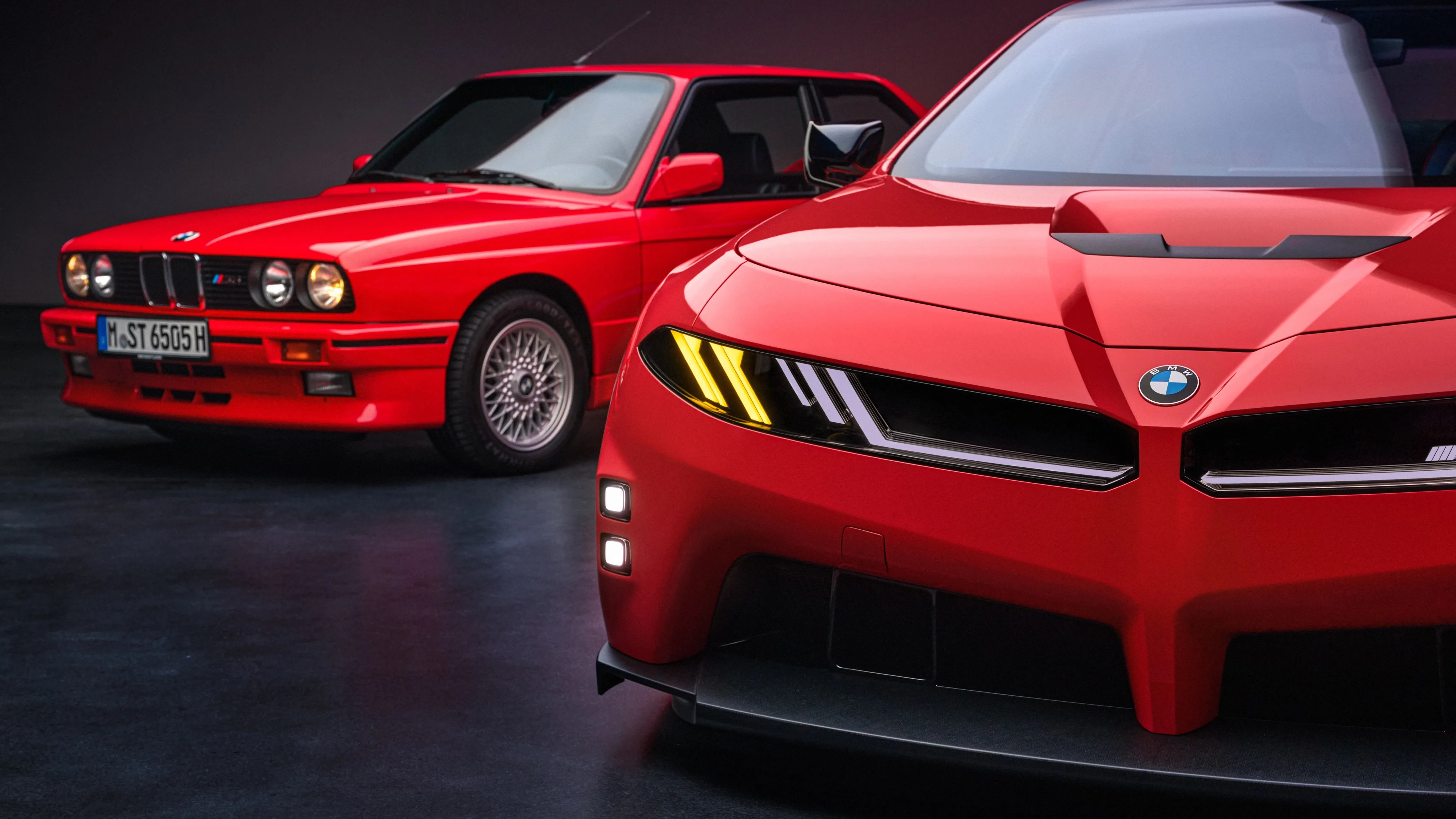 Front view of a red modern BMW with illuminated headlights next to a red classic BMW E30 M3 in a dark studio.
