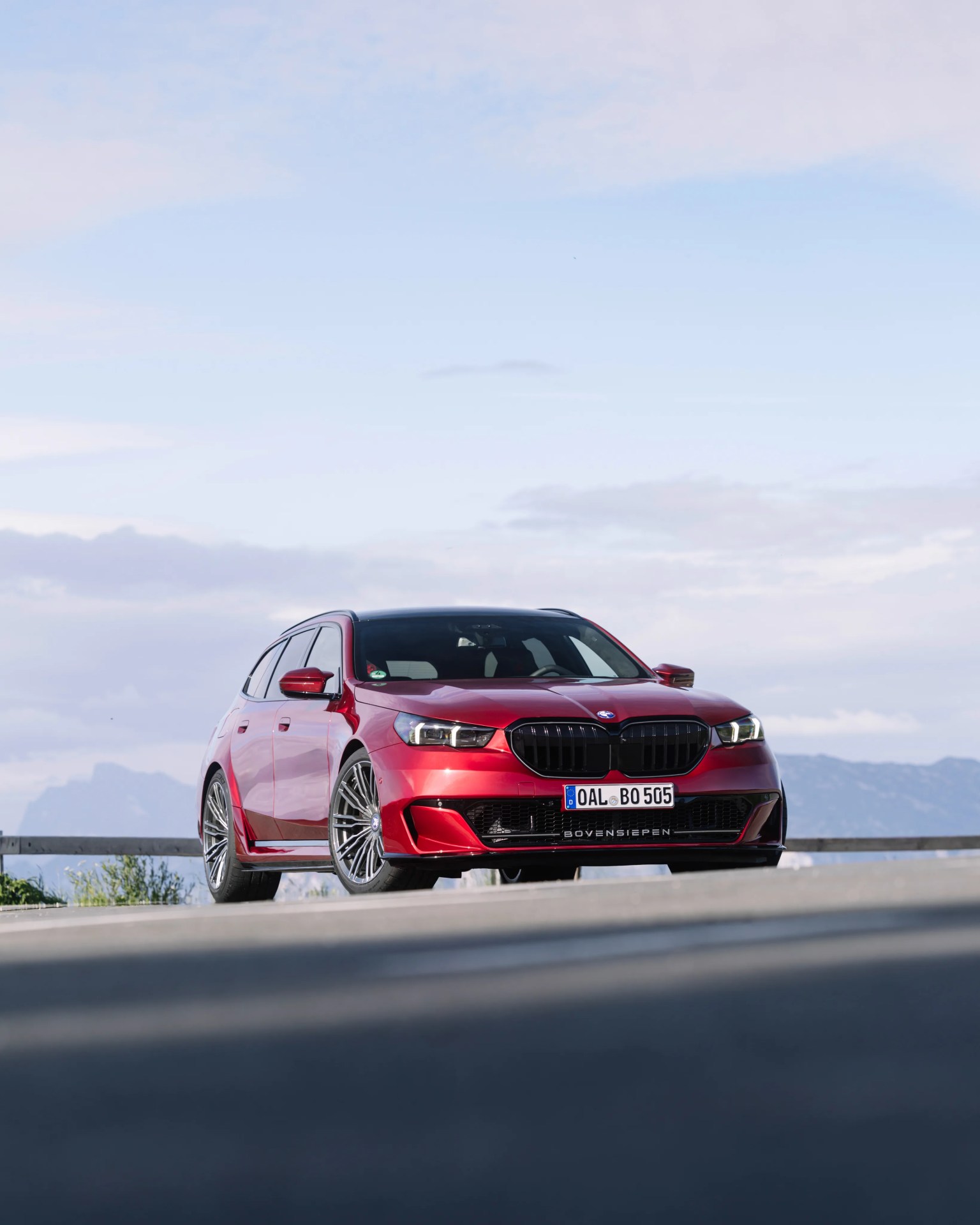 Red BMW station wagon with black grille and silver multi-spoke wheels on a road with mountains in the background.