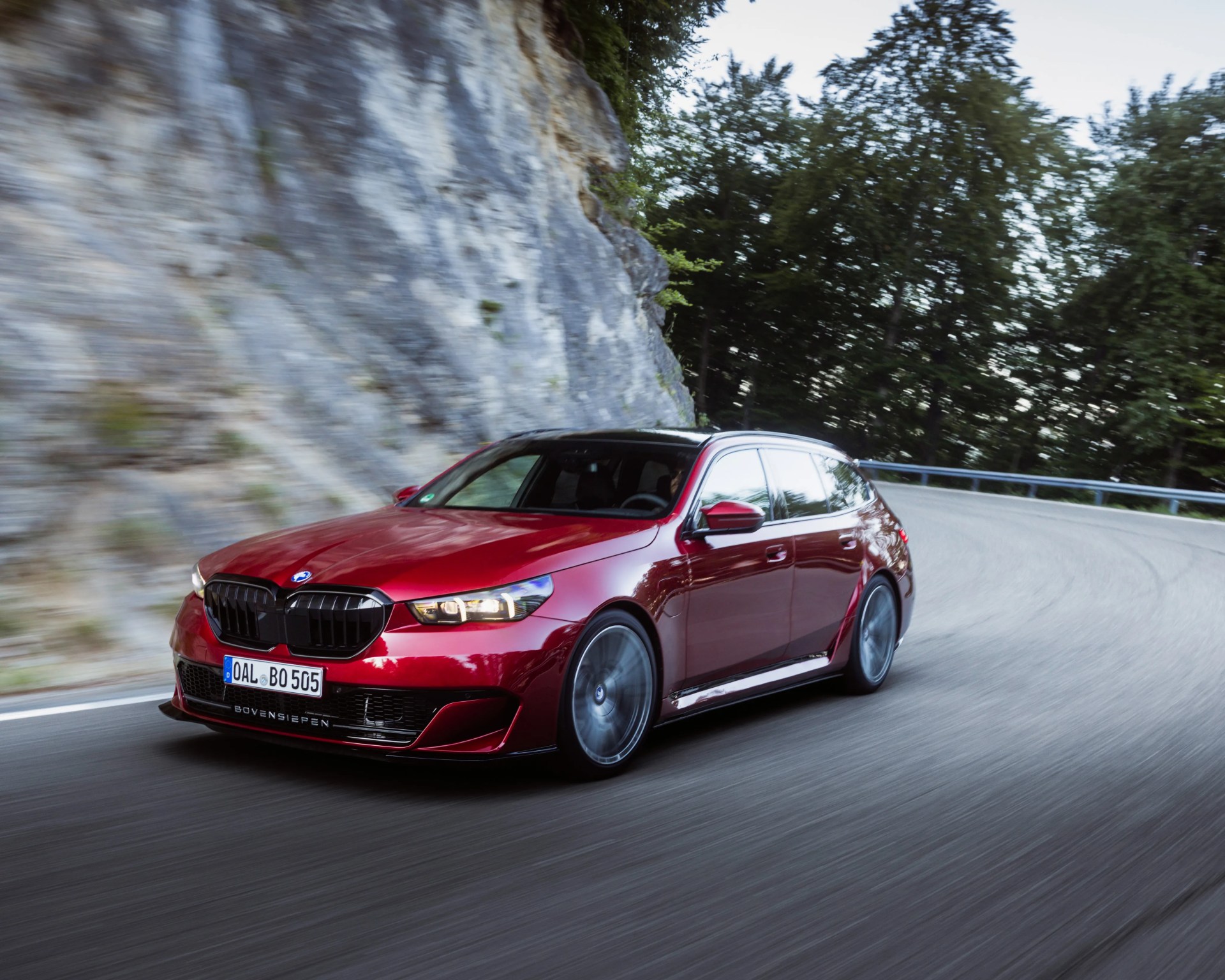 Red BMW station wagon with black roof driving on a winding mountain road with rocky cliff and trees.