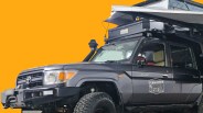 Black Toyota Land Cruiser with rooftop tent and off-road tires against an orange background.