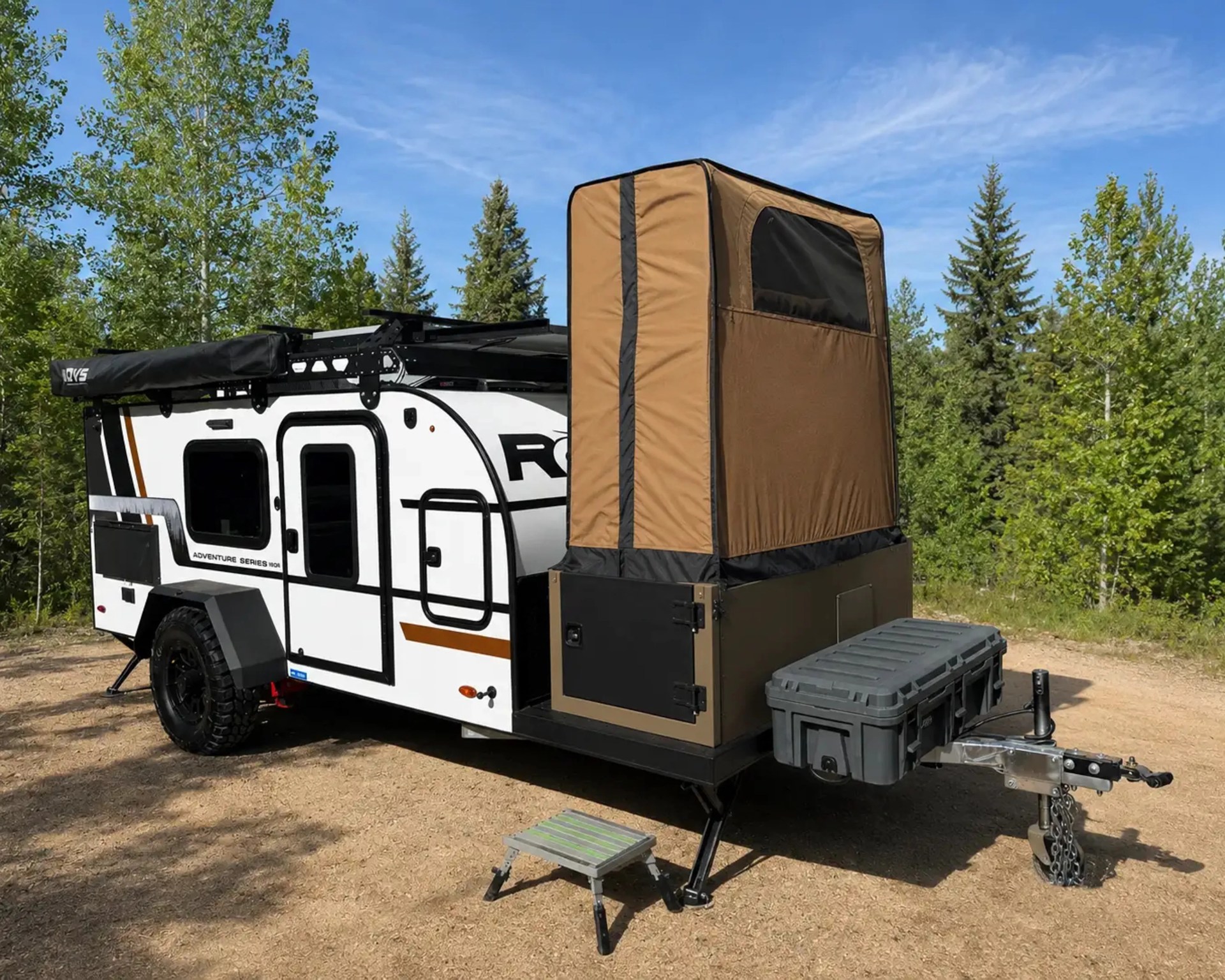 White off-road camper trailer with black trim and a tan rooftop tent extension, parked on a dirt surface with green trees in the background.