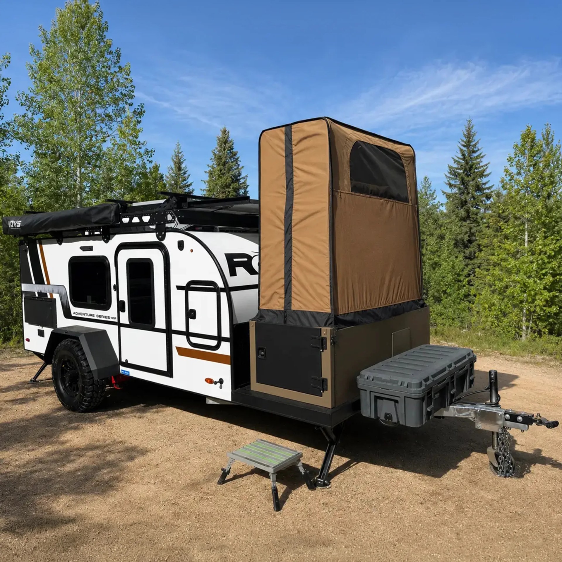 White and black off-road camper trailer with brown rooftop tent and gray storage box on a dirt ground surrounded by green trees.