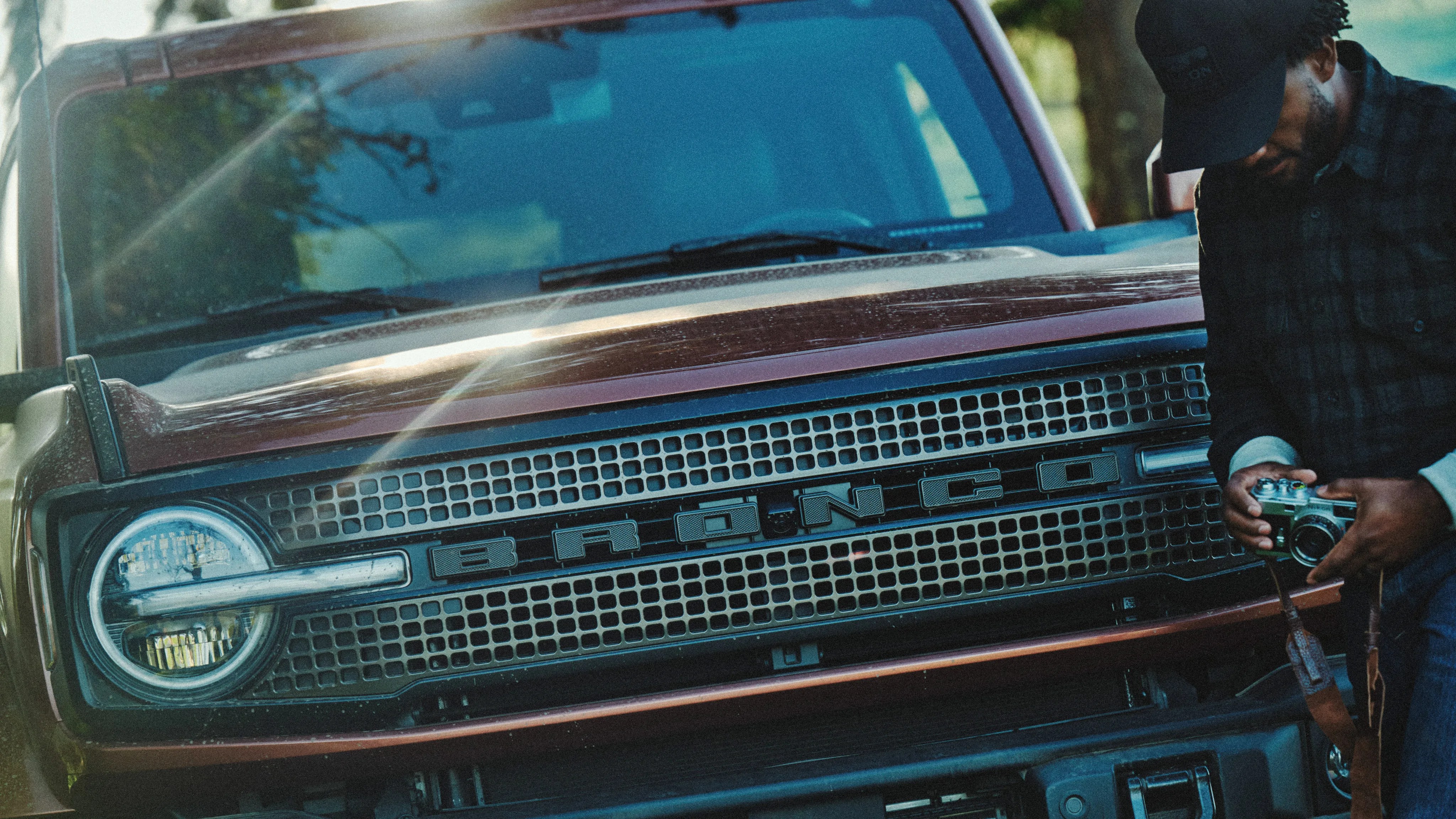 Front view of a brown Ford Bronco with a person holding a vintage camera on the right side.