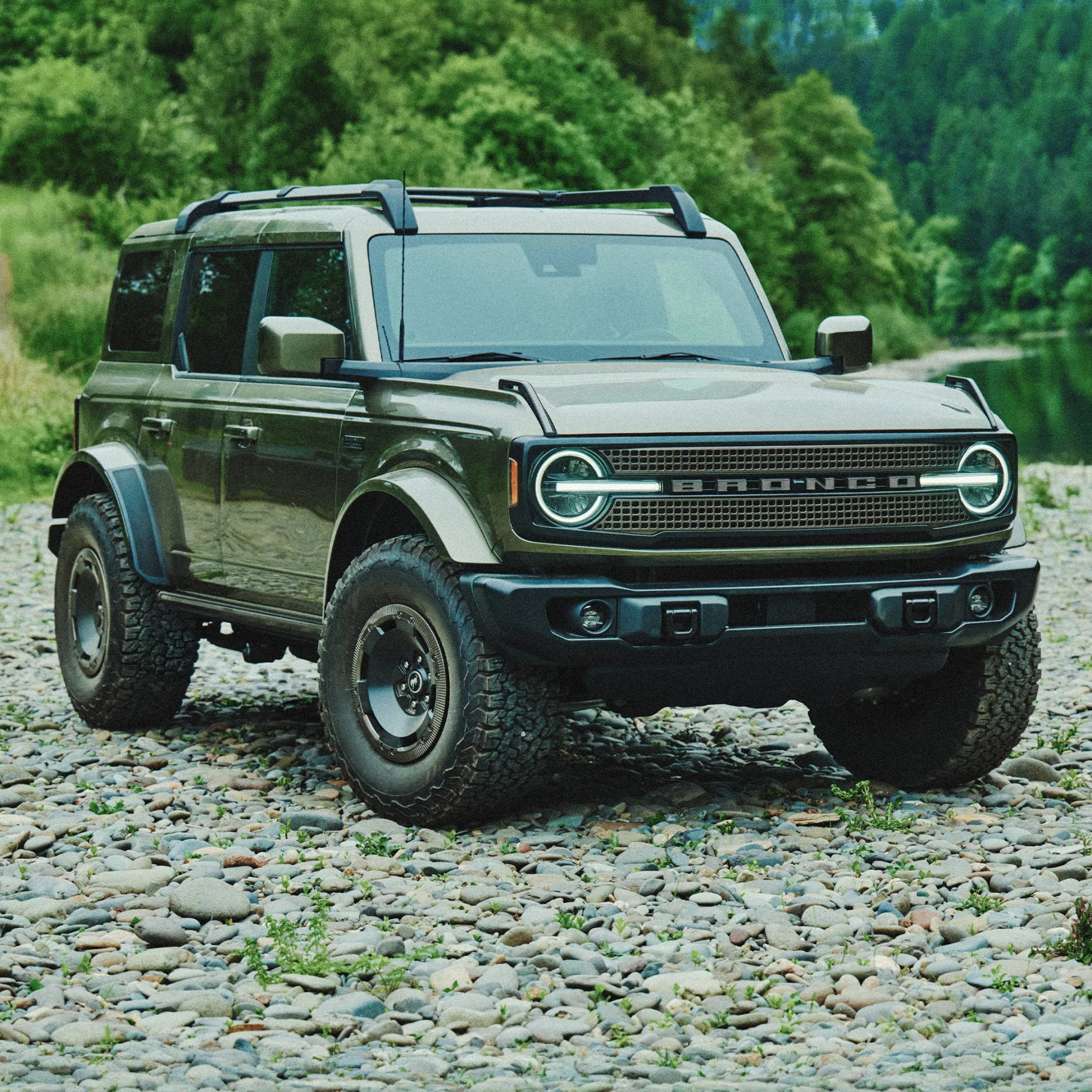 Green Ford Bronco SUV with roof rack parked on rocky ground near forest.