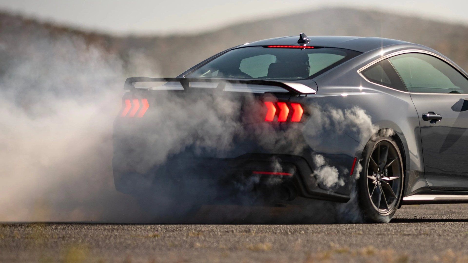 Dark gray sports car performing a burnout with smoke around the rear tires on a paved surface.