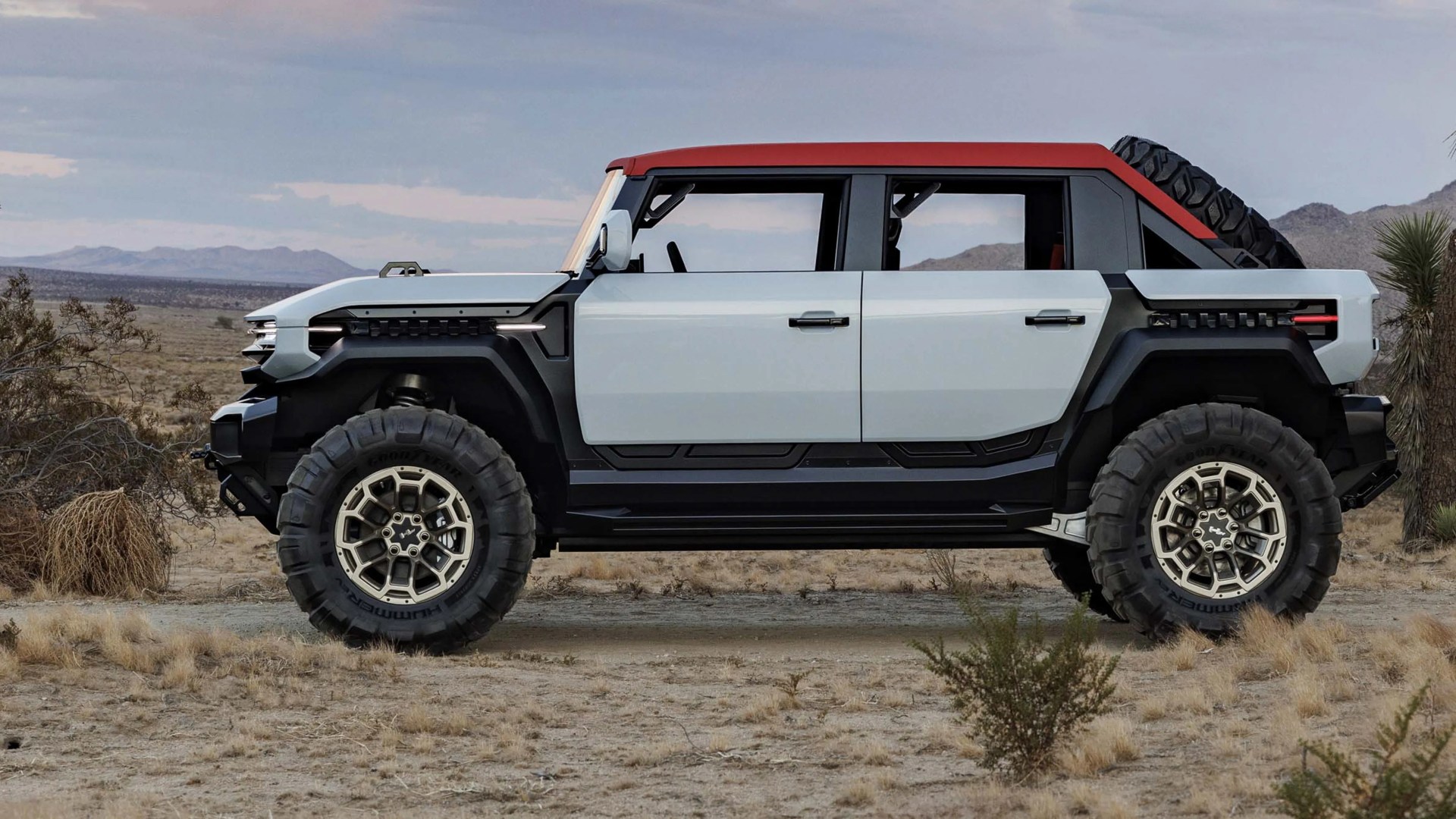 Off-road electric SUV with light gray body, red roof, large rugged tires, and spare tire mounted on rear.
