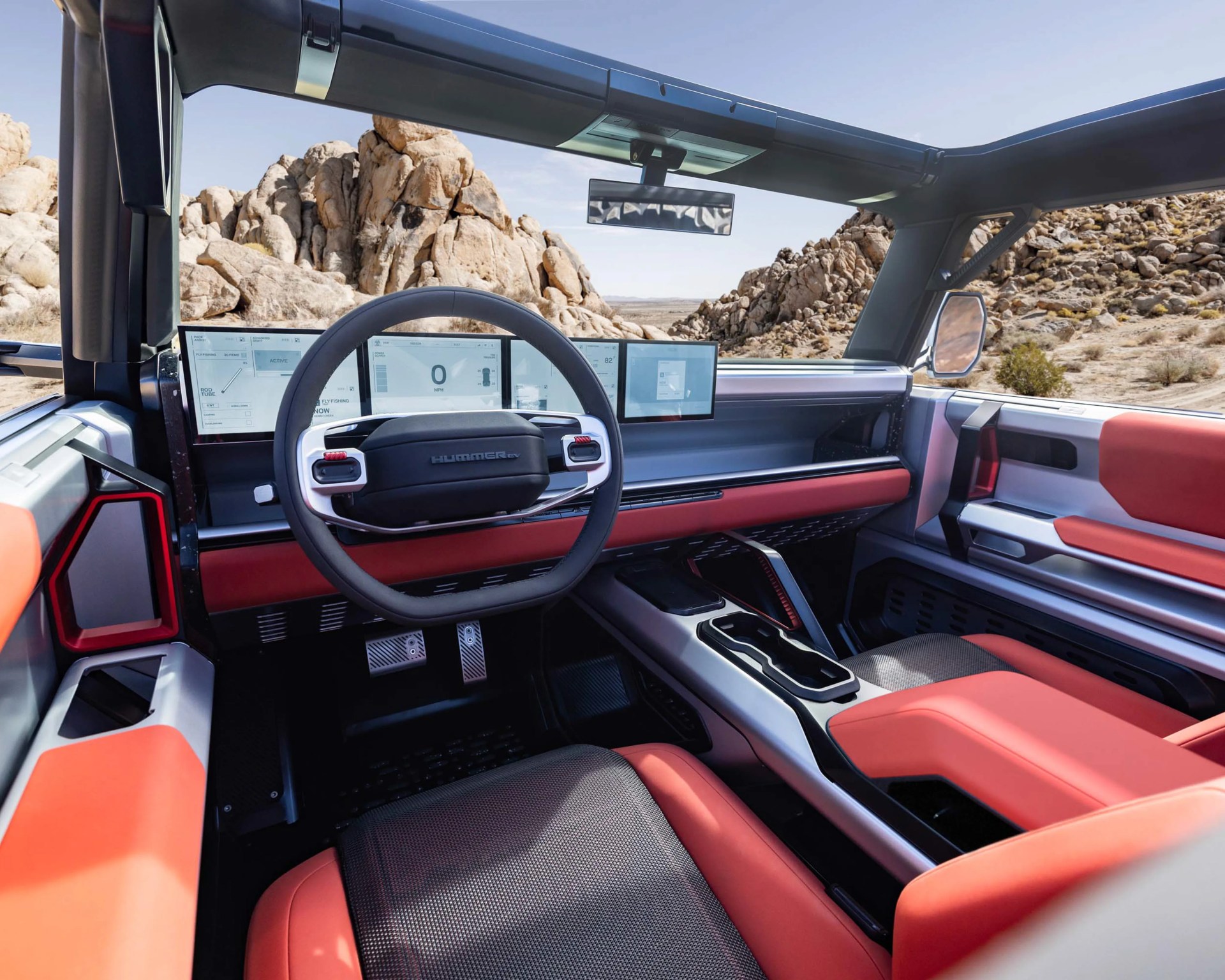 Interior of a Hummer EV with red and black seats, digital dashboard, and open roof showing rocky desert outside.