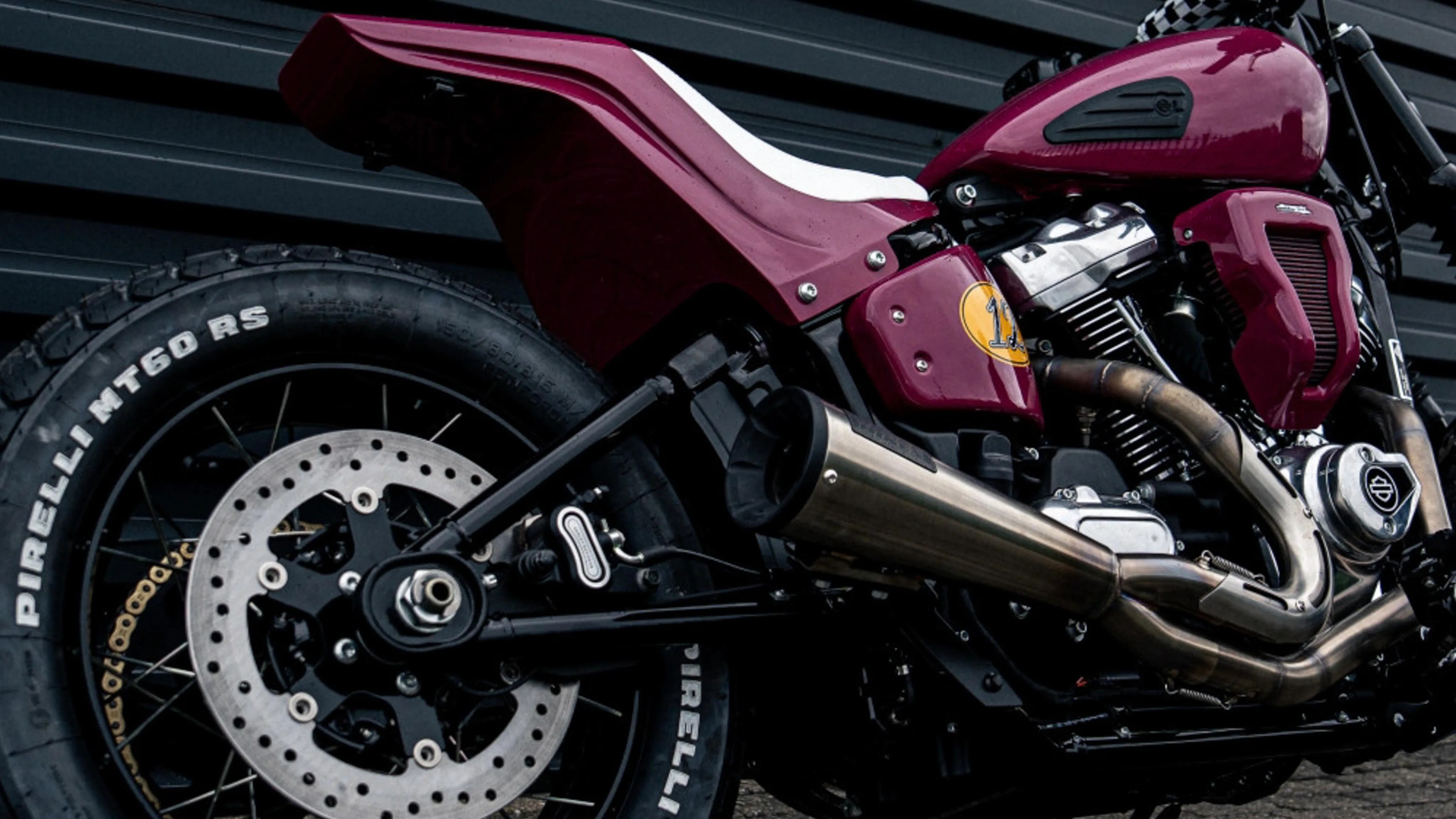 Close-up of a maroon motorcycle's rear wheel, exhaust pipes, and engine with Pirelli MT60 RS tire.