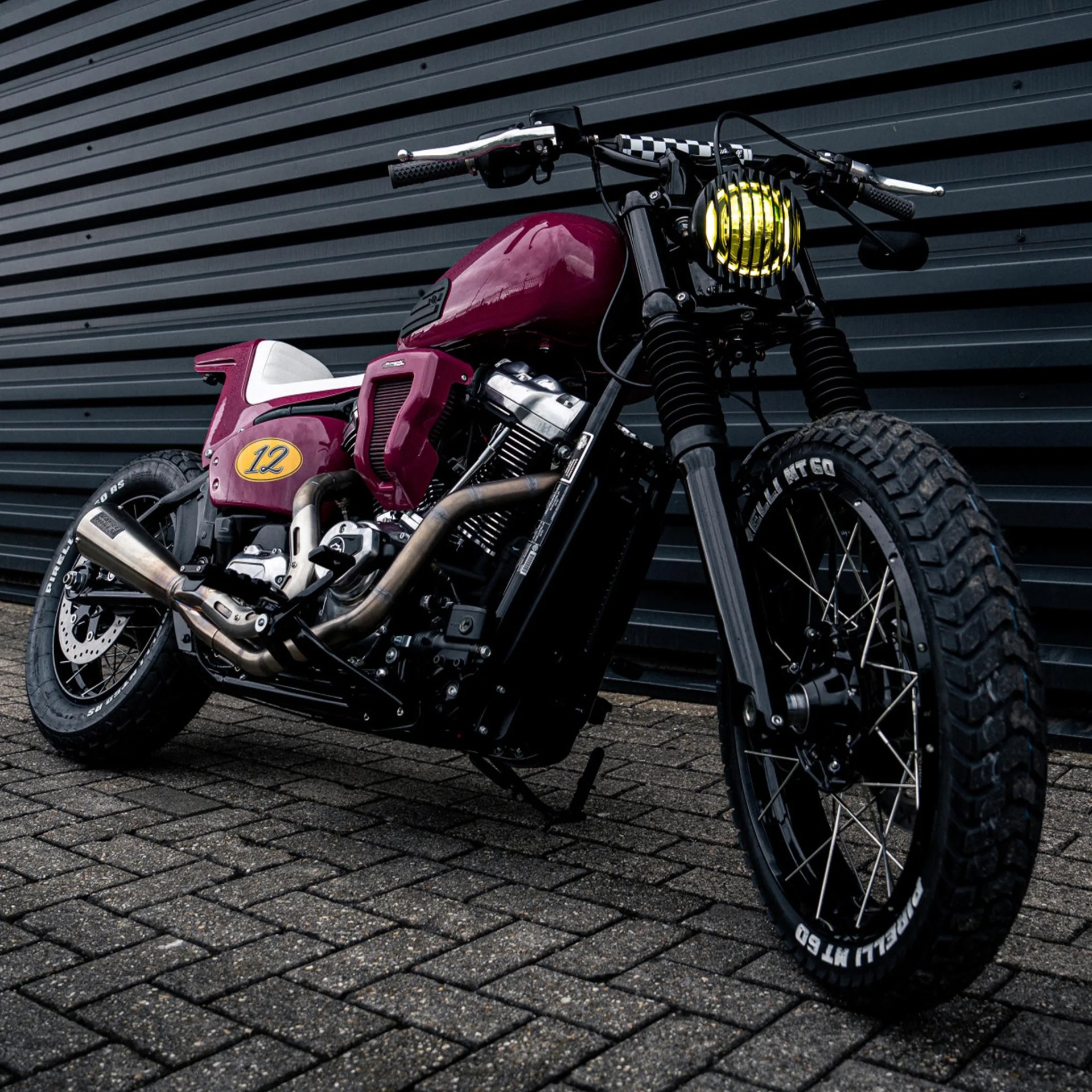 Custom maroon motorcycle with white seat, yellow headlight, and knobby Pirelli tires parked on brick pavement.