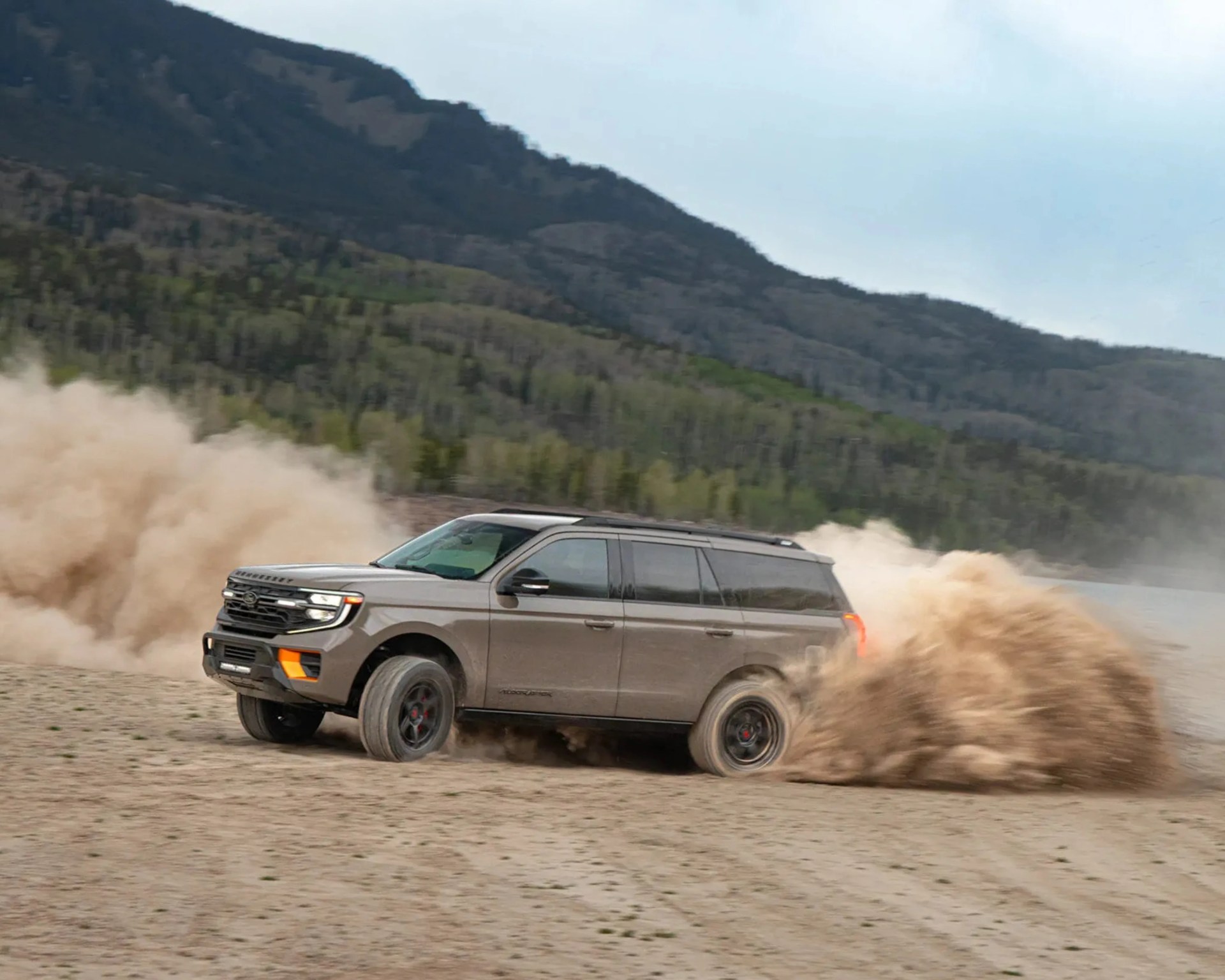 Gray Ford SUV kicking up dust while driving on a dirt road with forested hills in the background.