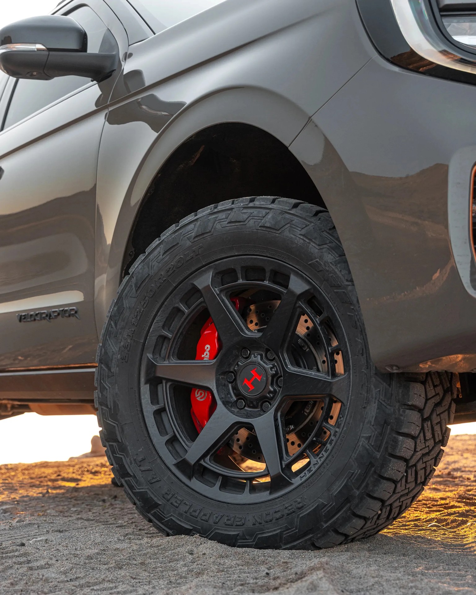 Black off-road tire with rugged tread and red brake caliper on a gray vehicle on sandy ground.