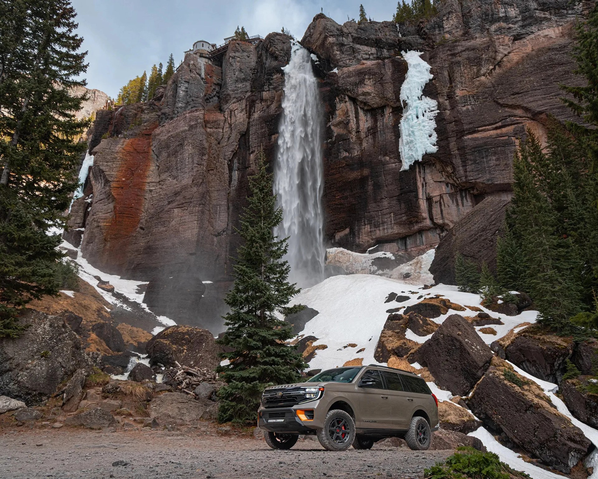 Brown SUV parked on rocky ground near a tall waterfall and snow-covered cliffs with evergreen trees.