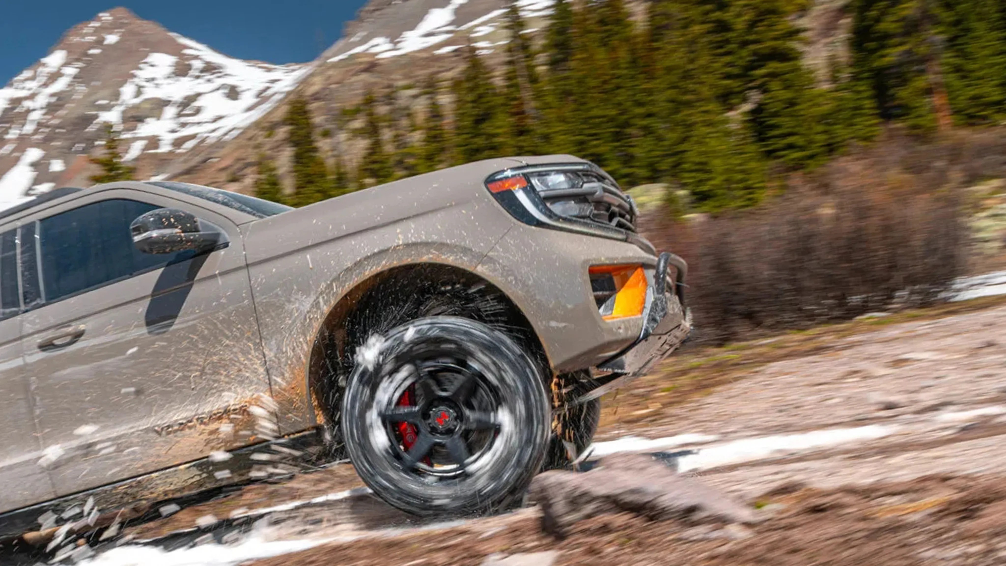 Beige off-road truck with black wheels splashing mud on a snowy rocky trail with pine trees and snow-capped mountains in the background.