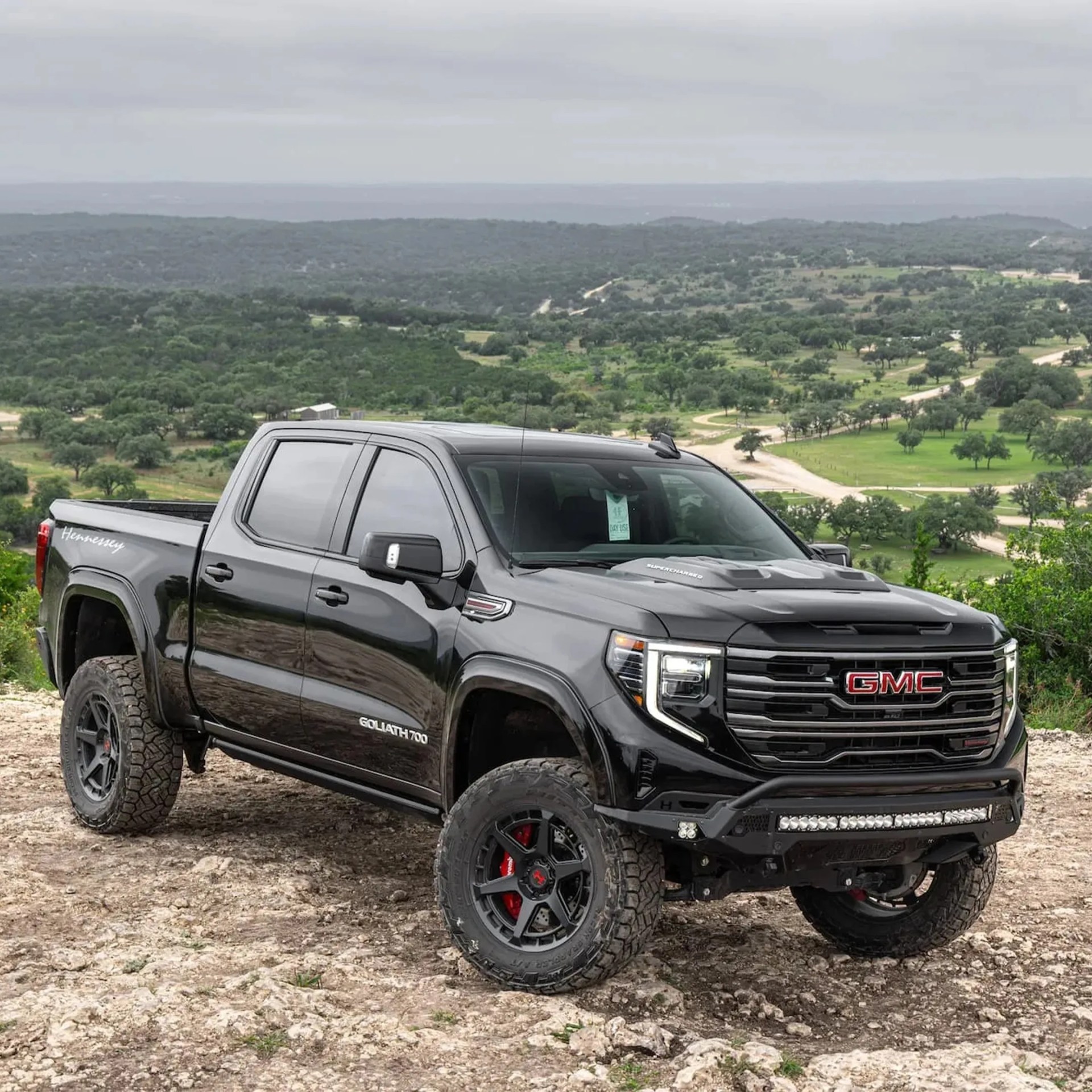 Black GMC Goliath 700 pickup truck with off-road tires parked on rocky terrain with green hills in the background.