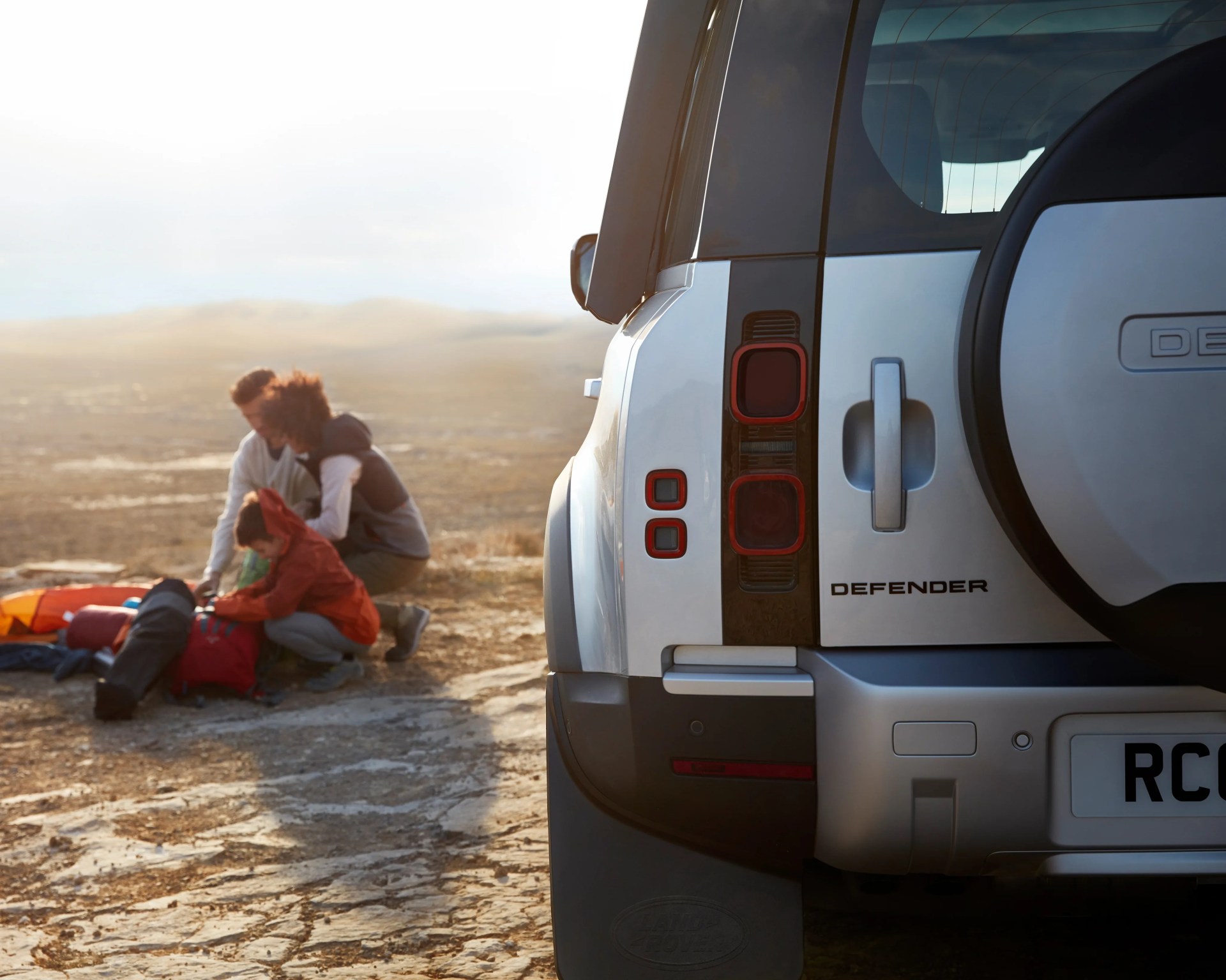 Rear view of a silver Land Rover Defender with a spare tire on the back and people packing gear in the background on a rocky terrain.
