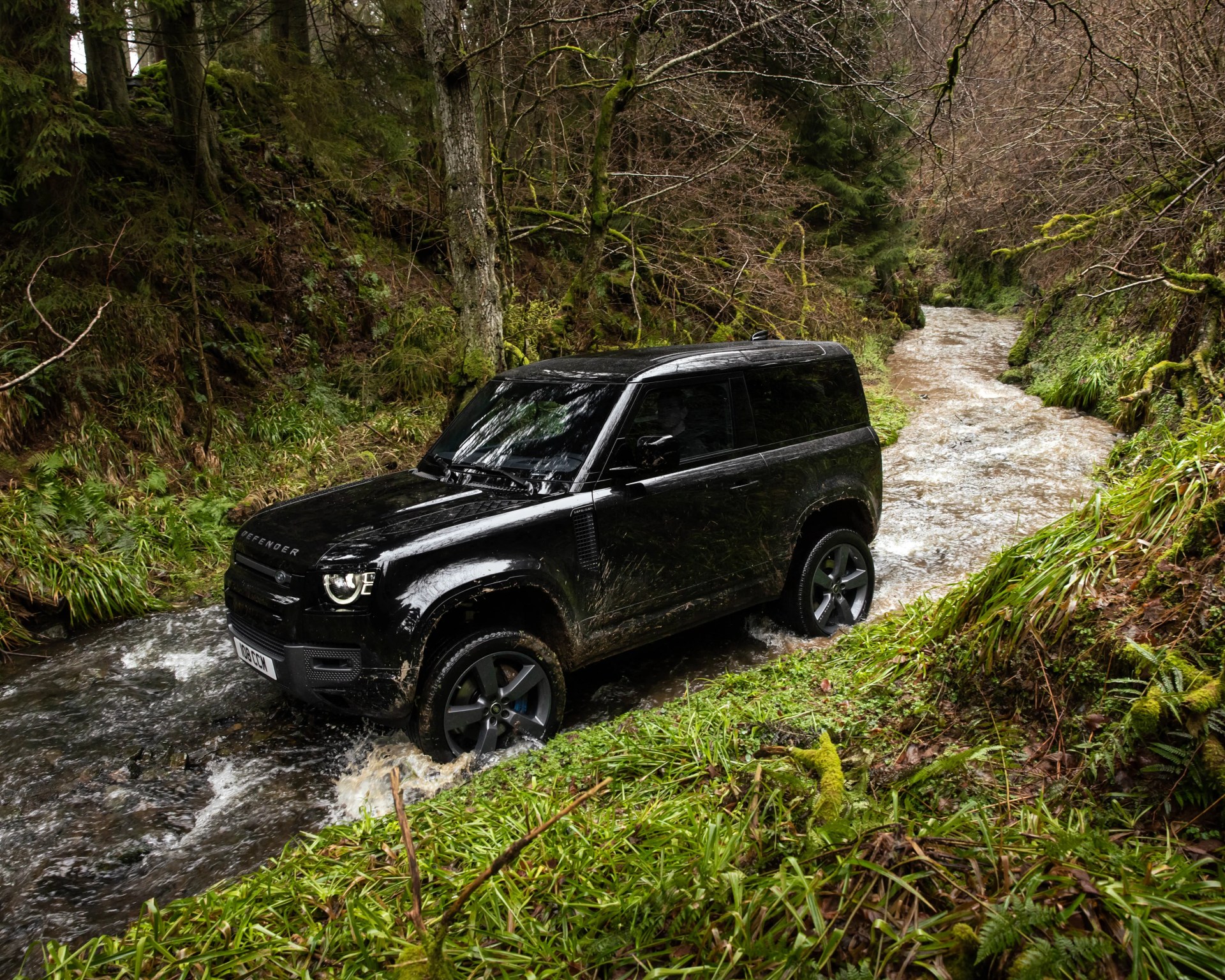 Black Land Rover Defender driving through a shallow forest stream surrounded by mossy trees and green grass.