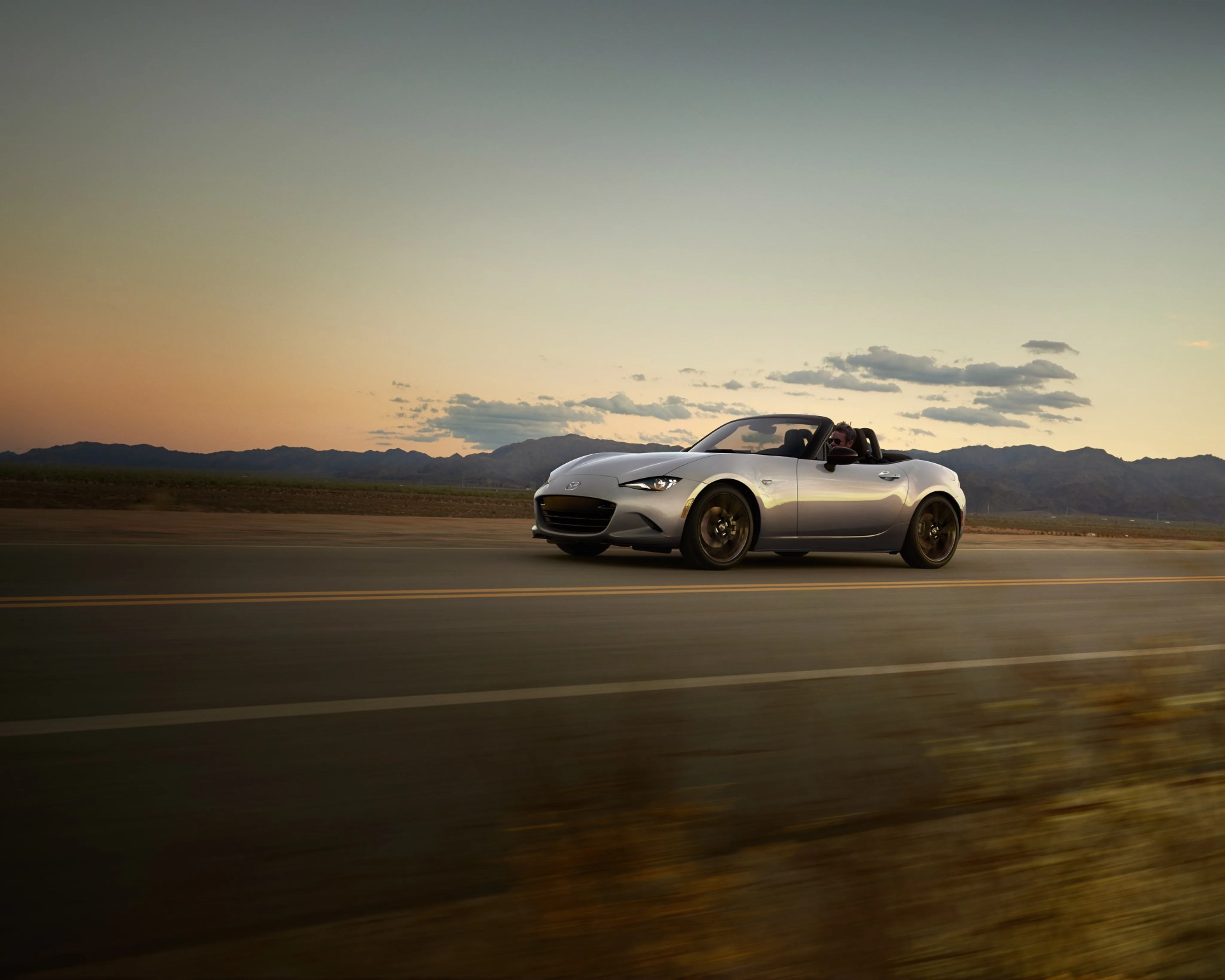 Silver convertible sports car driving on a road at sunset with mountains in the background.