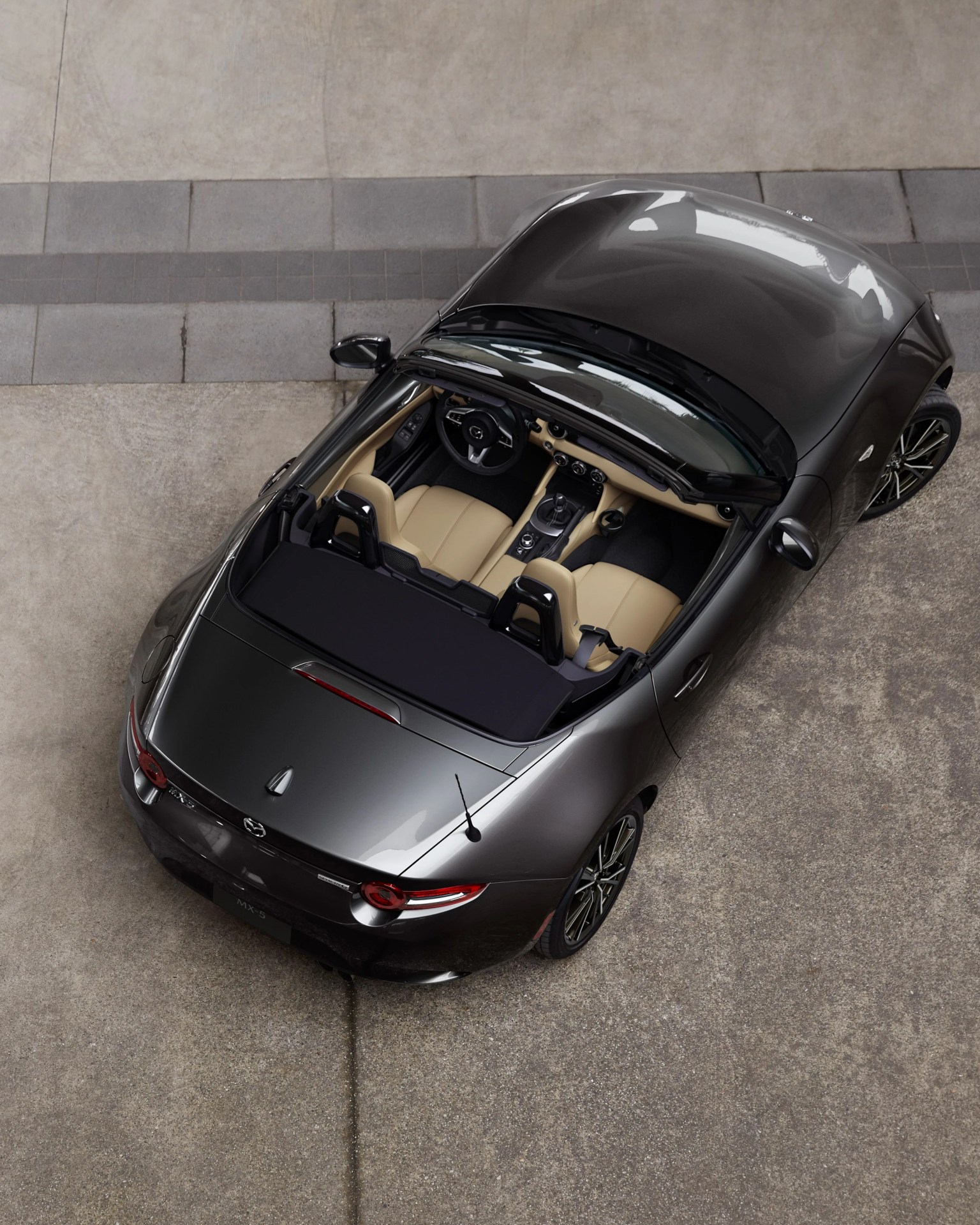Dark gray Mazda MX-5 convertible with beige leather seats viewed from above on a concrete surface.