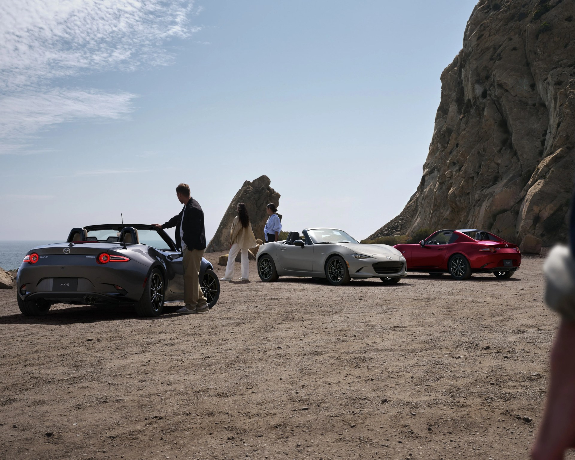 Three Mazda MX-5 convertible sports cars in gray, silver, and red parked on a rocky cliffside with people nearby.