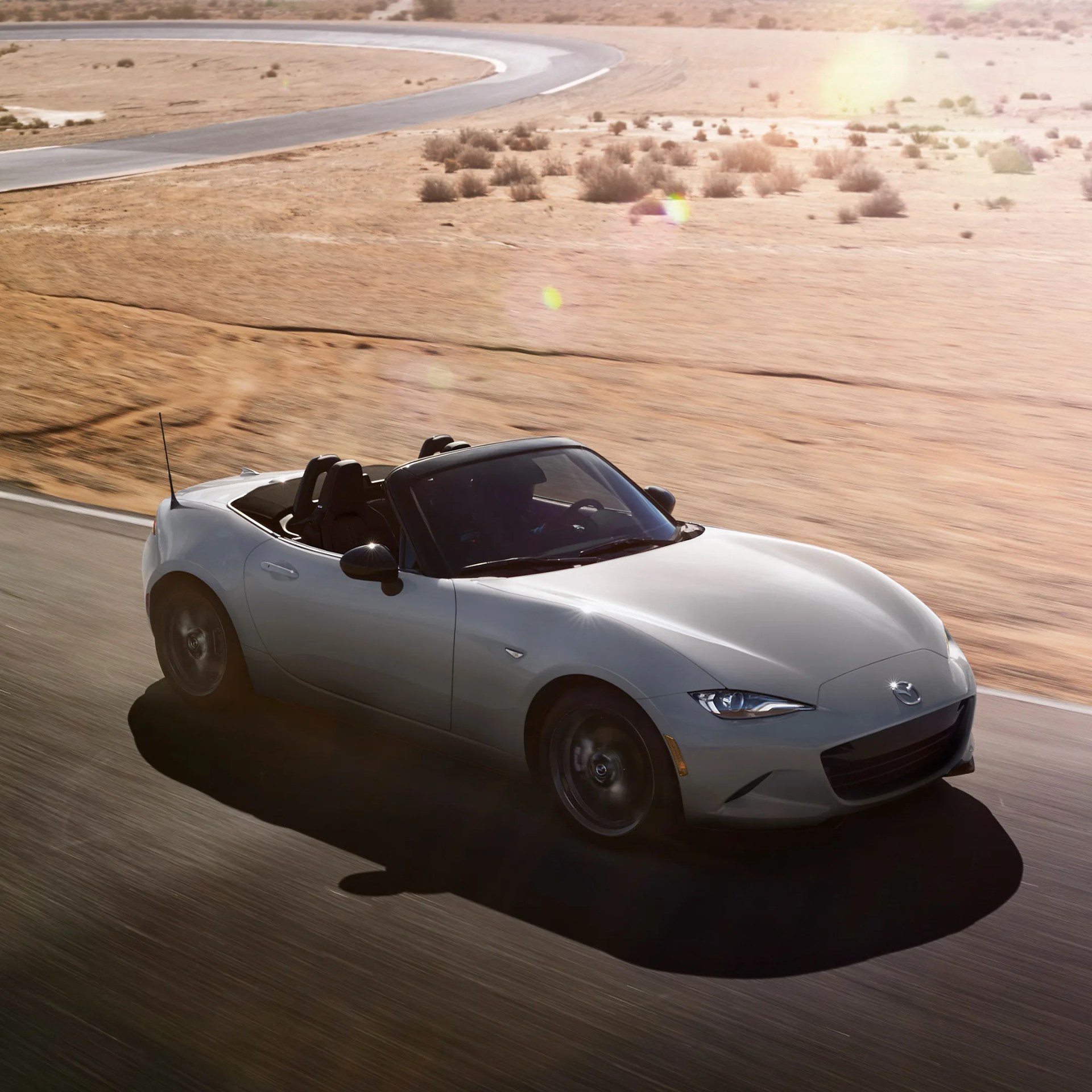 Silver Mazda convertible sports car driving on a desert racetrack with sunlight glare.