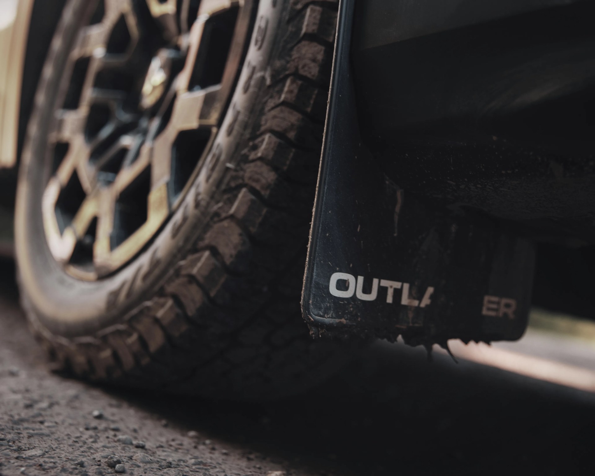 Close-up of a rugged off-road tire and a black mudflap with partially visible "OUTLA" text on a dirt surface.
