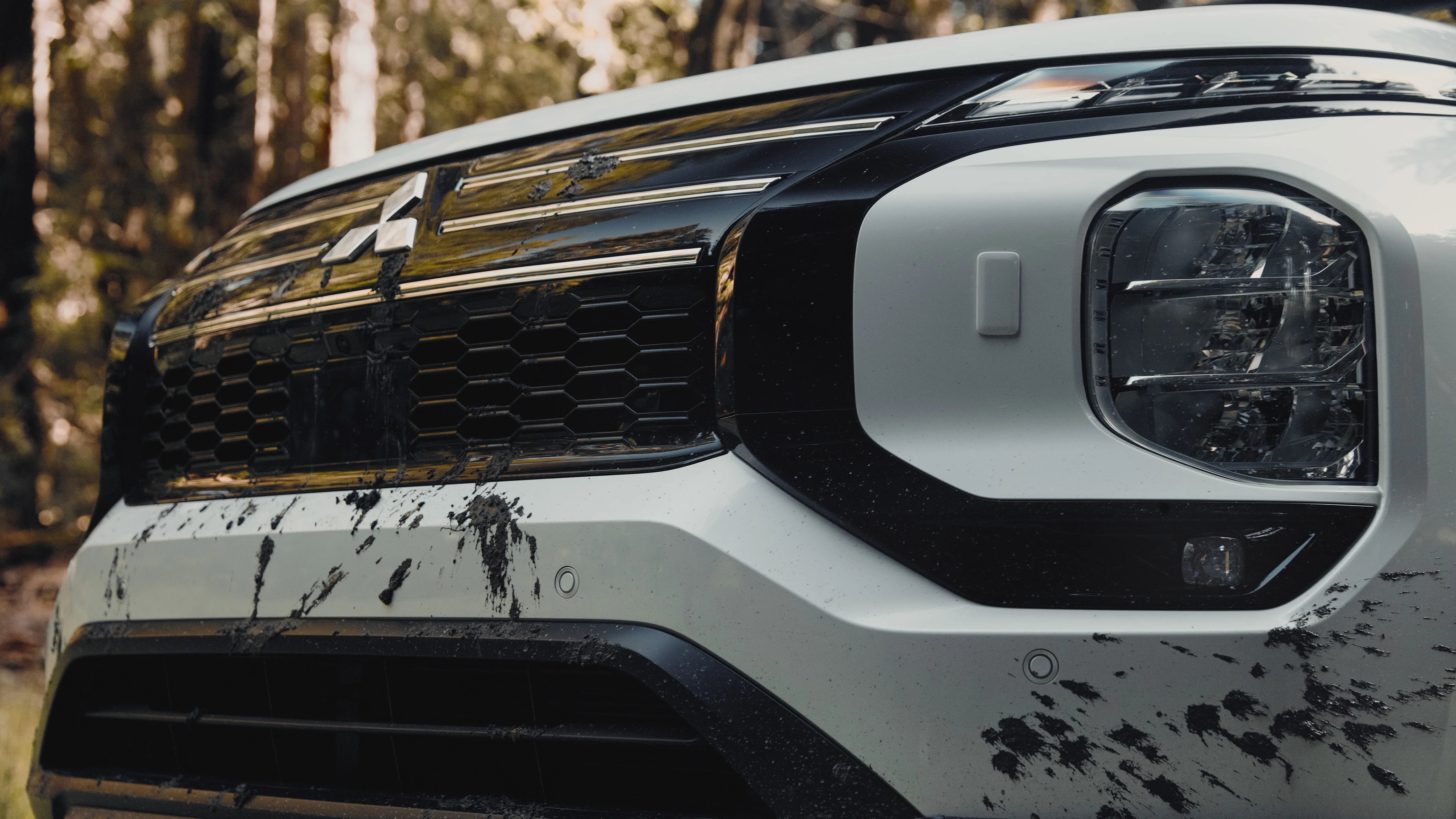 Front grille and headlight of a white Mitsubishi vehicle with mud splatters on the bumper and grille.