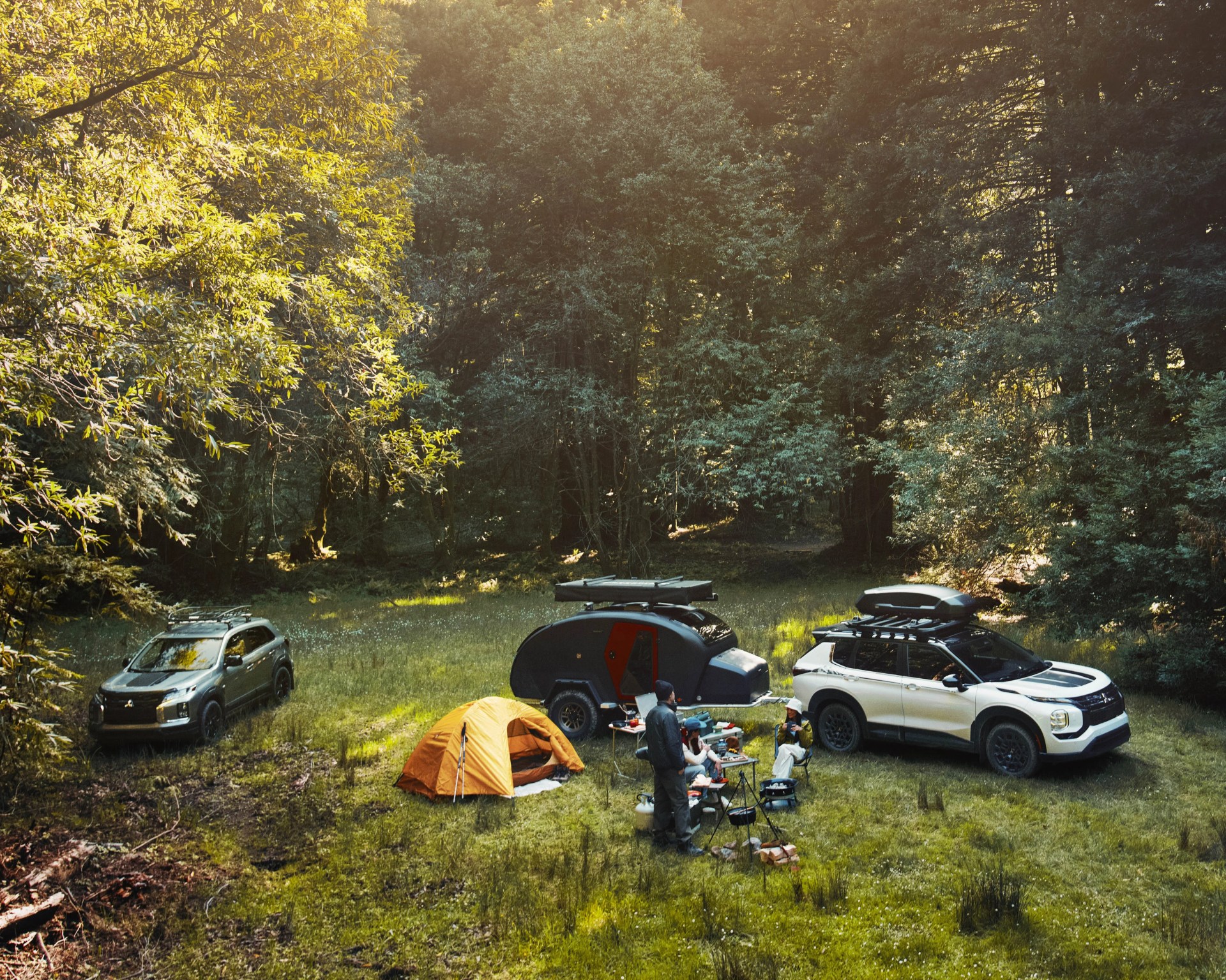 Orange camping tent pitched on grass near two SUVs and a small black and red camper trailer in a forest clearing.