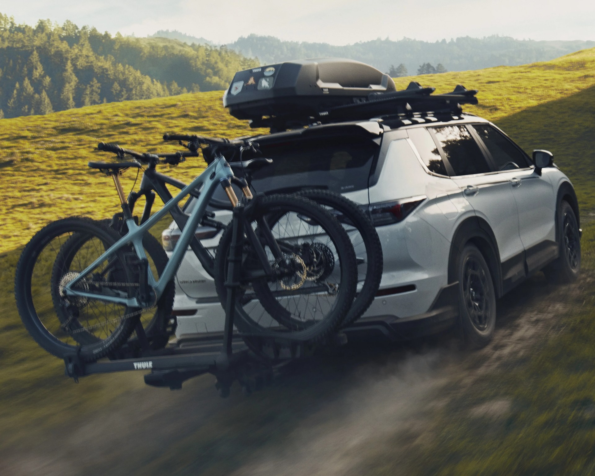 White SUV with roof cargo box and two mountain bikes mounted on a rear bike rack driving on a dirt road in a grassy hilly area.