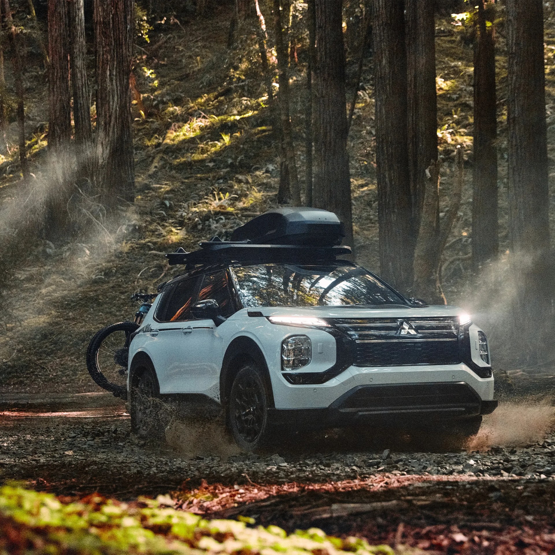 White Mitsubishi SUV with roof cargo box and rear-mounted bicycle driving on a forest trail.