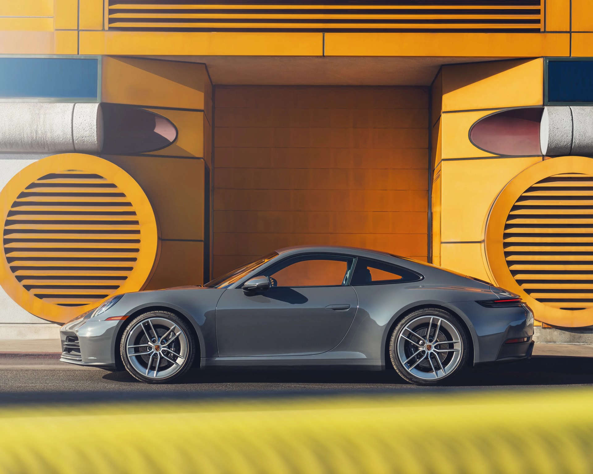 Gray Porsche sports car parked in front of a yellow and orange industrial wall with circular vents.