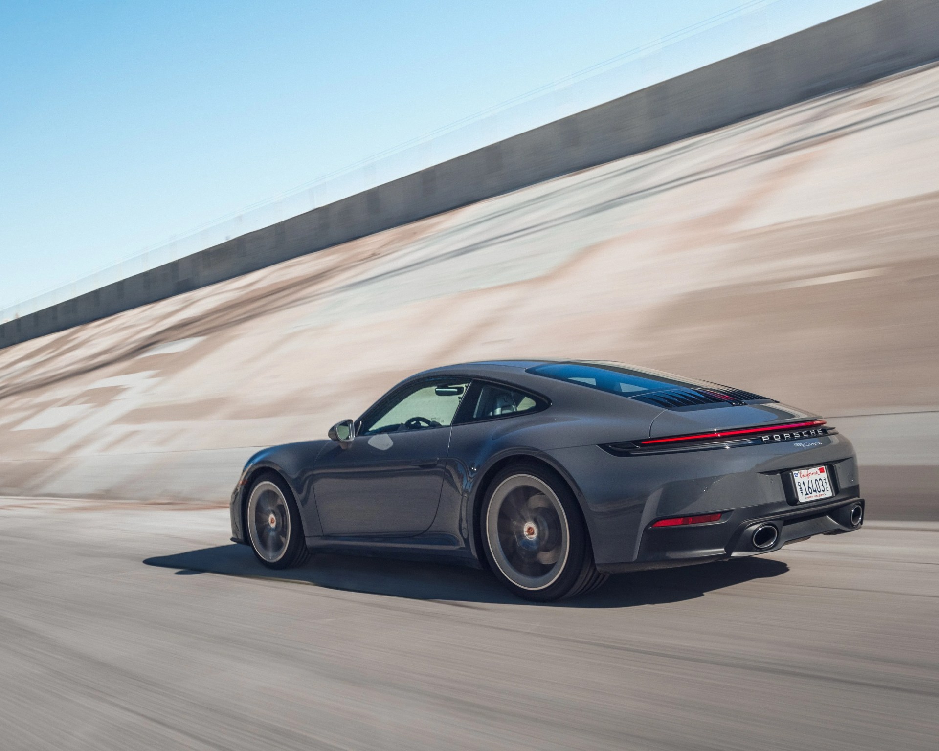 Dark gray Porsche 911 Carrera driving on a concrete road with a sloped wall in the background.
