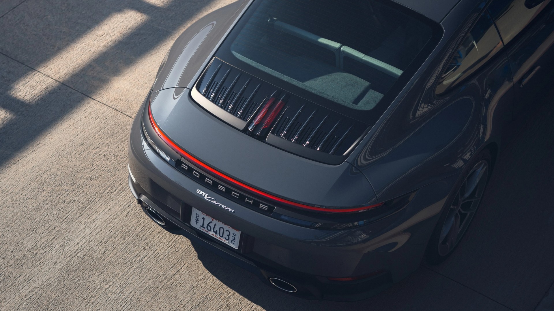 Rear view of a gray Porsche 911 Carrera with illuminated rear light strip and visible license plate on concrete ground.