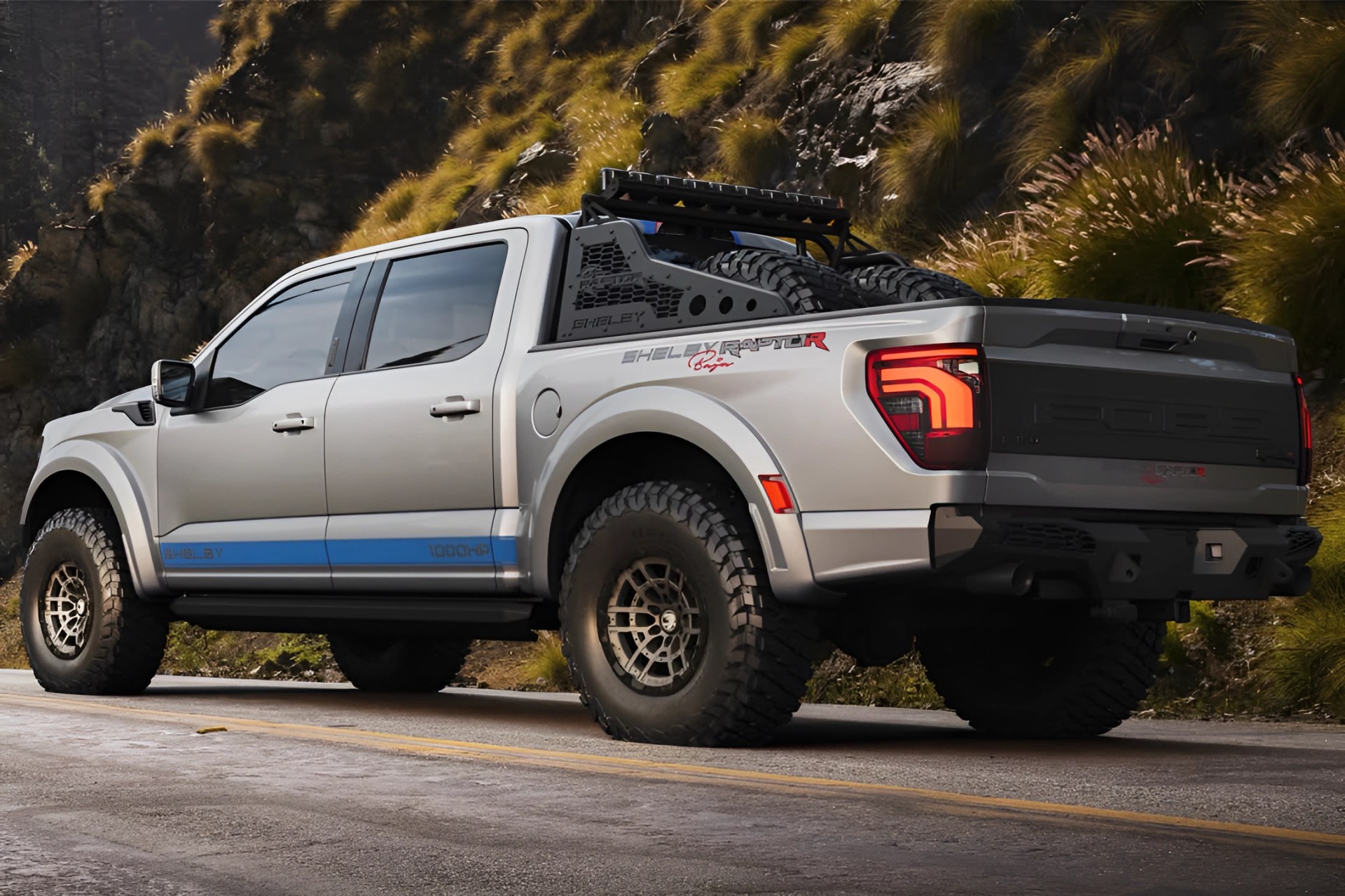Silver Shelby Raptor pickup truck with off-road tires and spare tires in the bed, parked on a road with rocky hillside background.