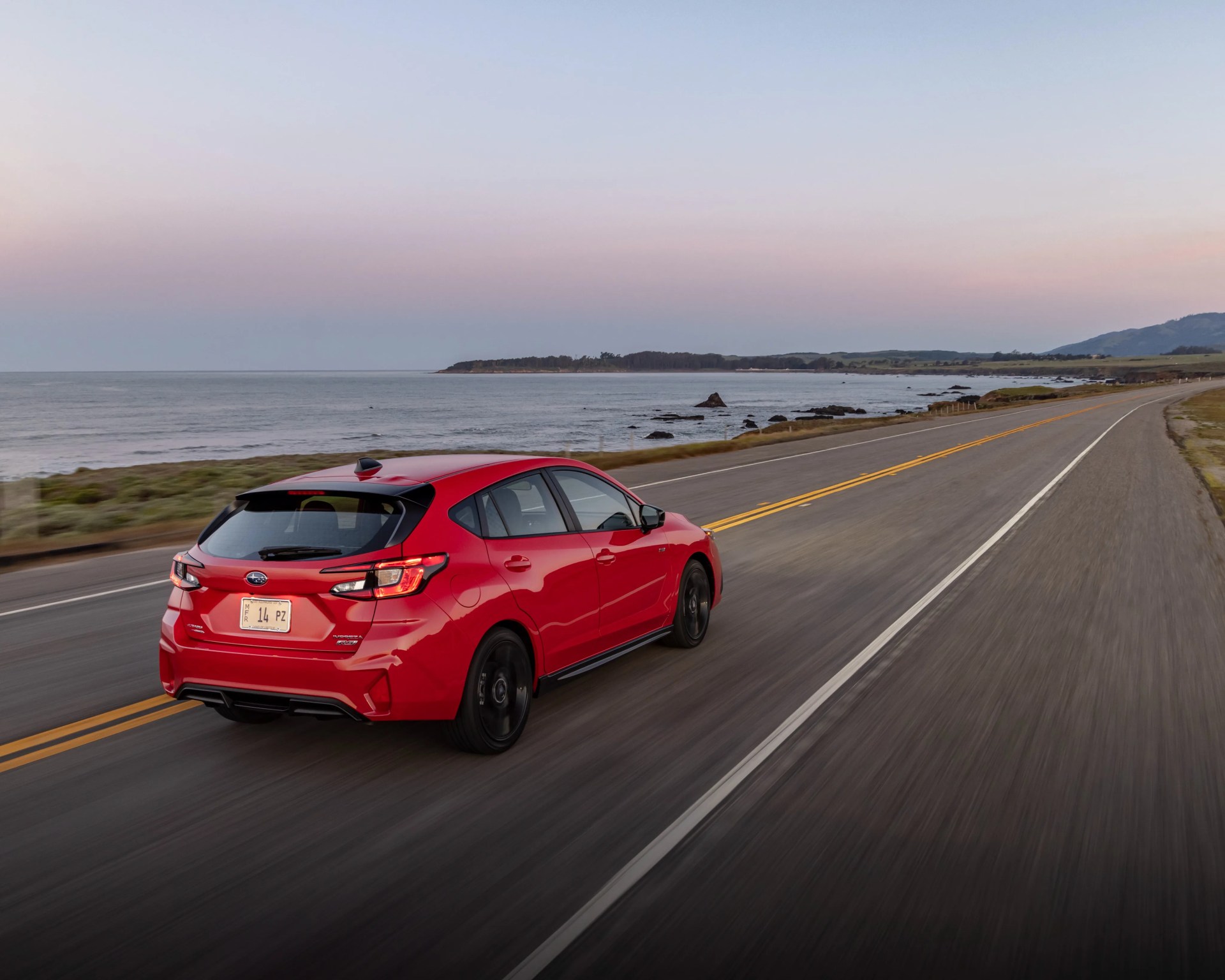 Red Subaru hatchback driving on a coastal highway at dusk with ocean and hills in the background.