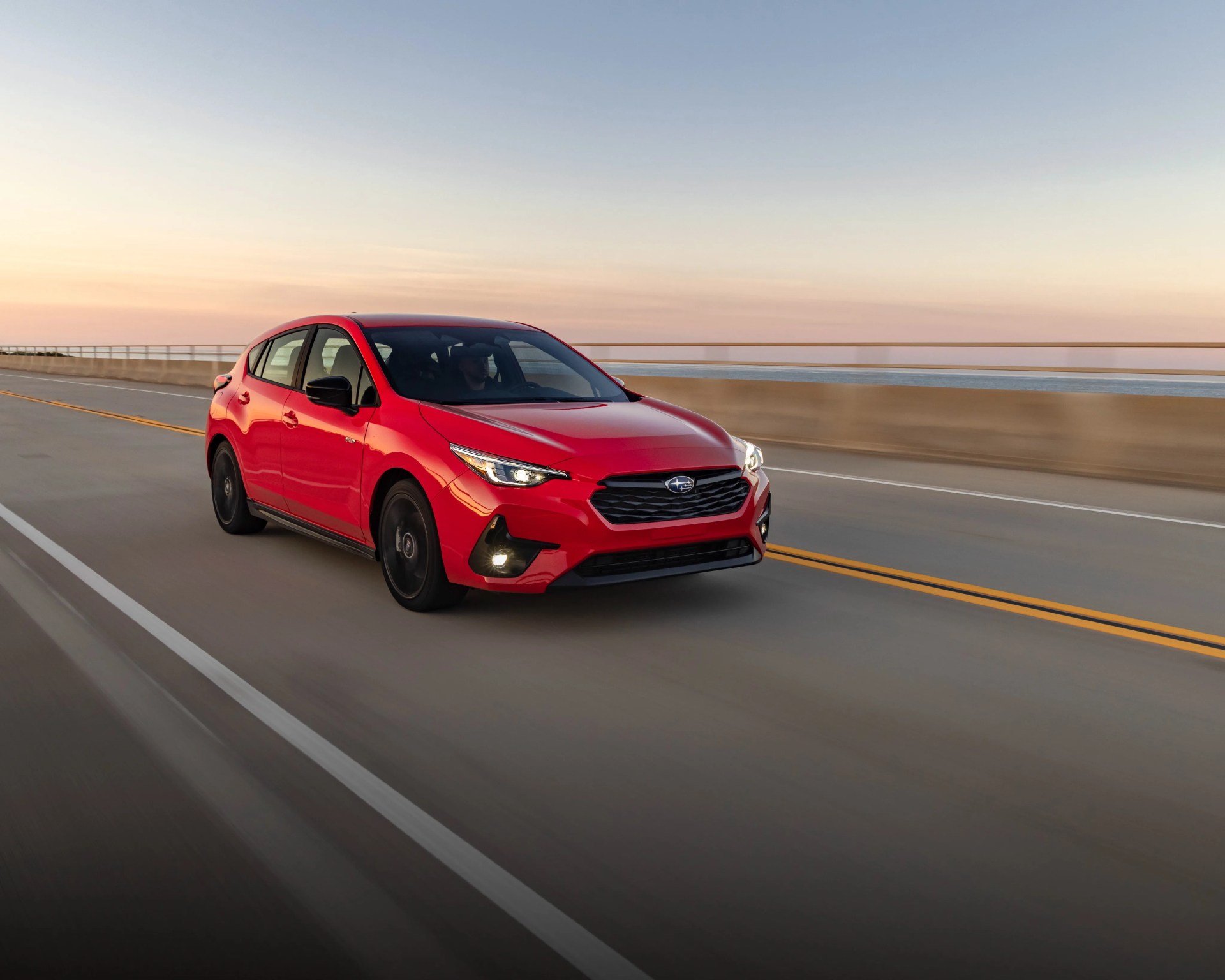 Red Subaru hatchback driving on a coastal highway at sunset.