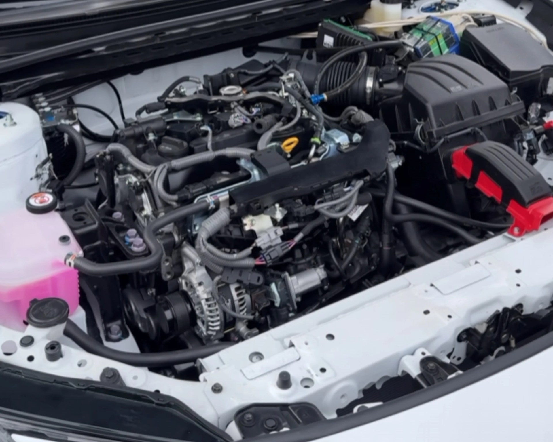Car engine compartment with visible alternator, air intake, wiring, and coolant reservoir with pink fluid.