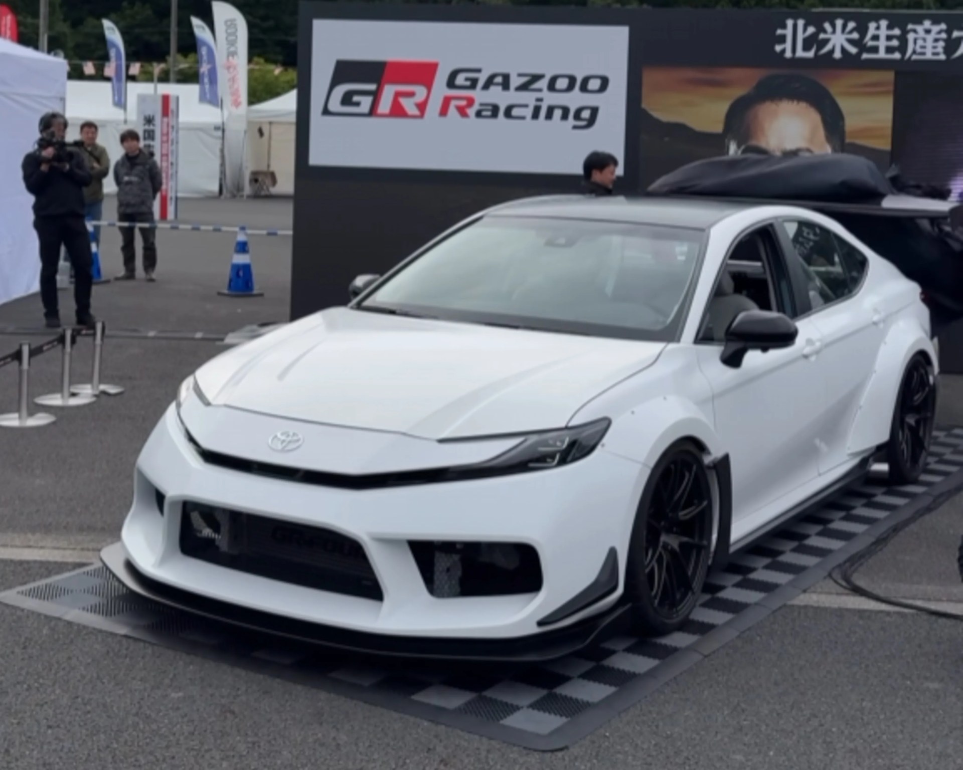 White Toyota sedan with black roof and sporty body kit displayed outdoors near a Gazoo Racing sign.