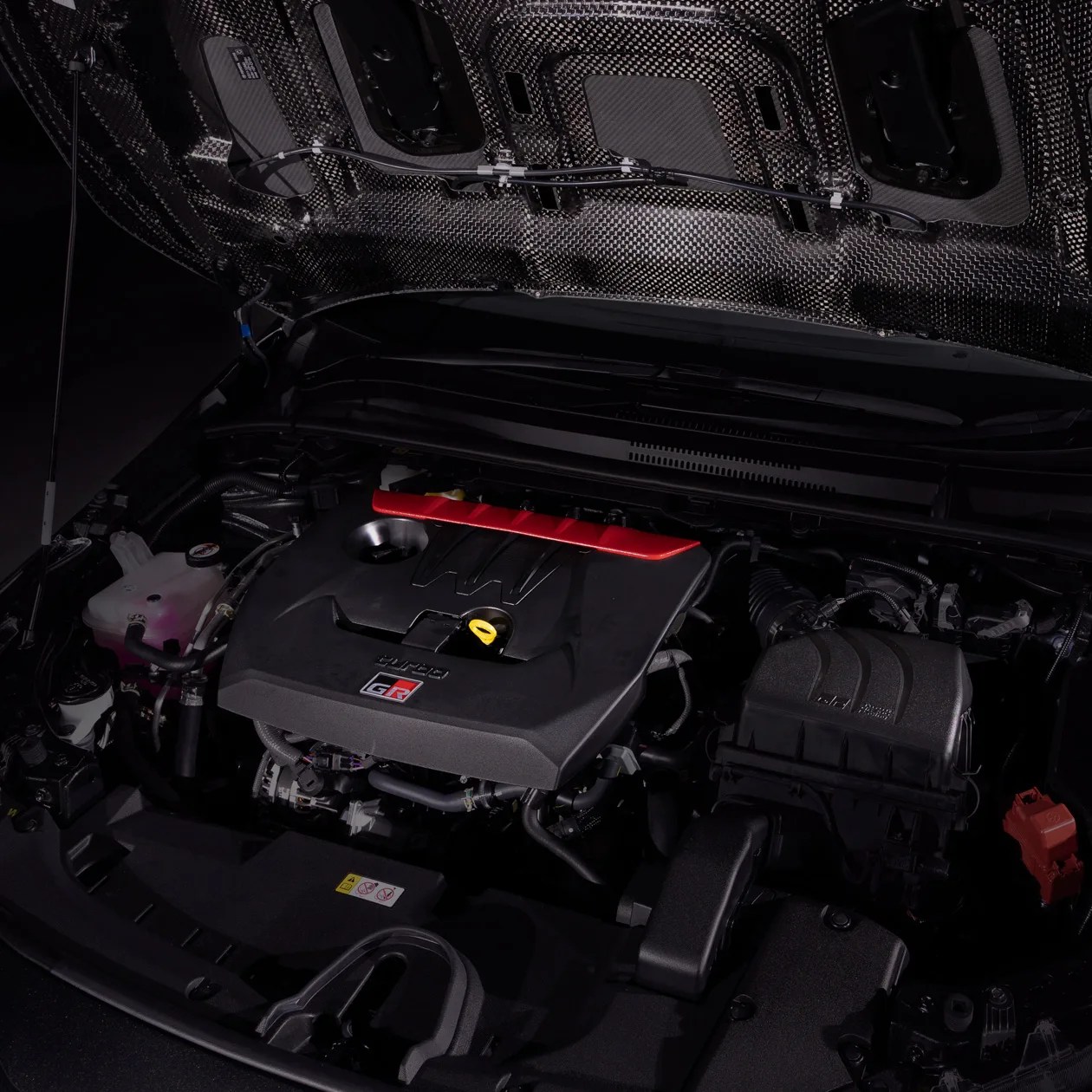 Car engine bay with a black engine cover featuring a red accent and a "GR" logo, and a carbon fiber hood interior.