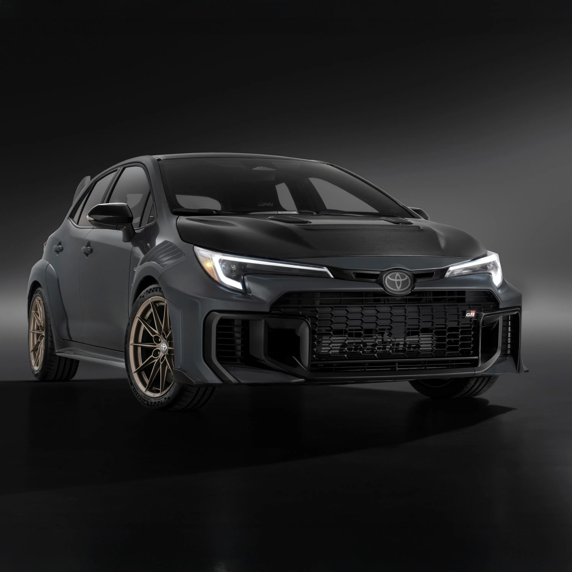 Matte black Toyota GR hatchback with bronze alloy wheels and aggressive front grille on a dark background.
