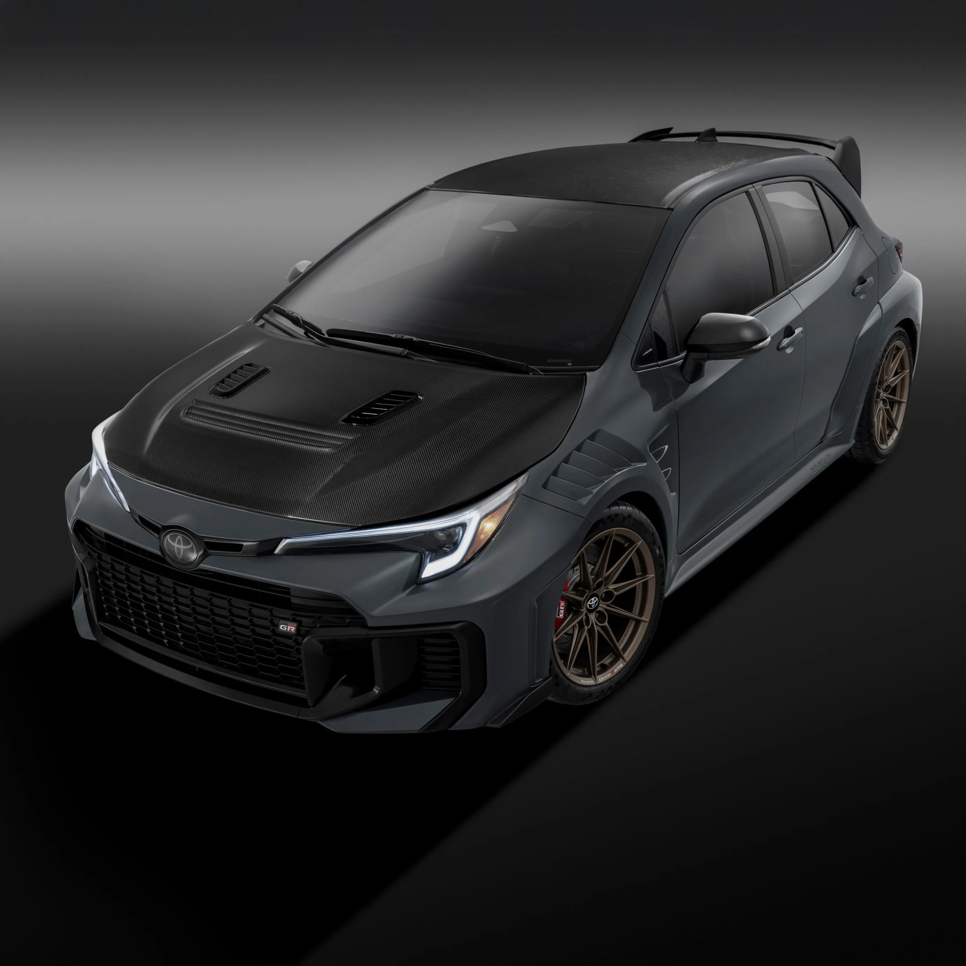 Matte gray Toyota GR hatchback with carbon fiber hood and roof, bronze wheels, and rear spoiler.