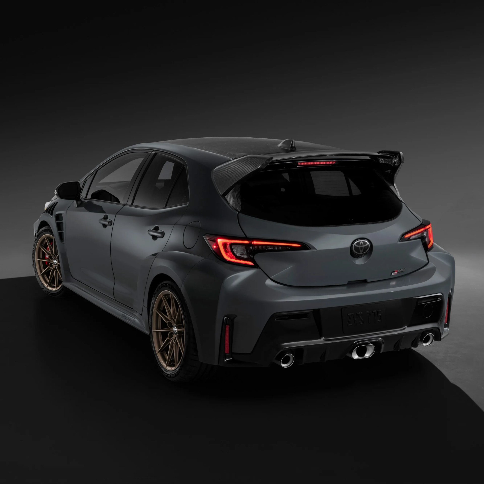Rear side view of a gray Toyota GR Yaris with bronze wheels and carbon fiber rear spoiler against a dark background.