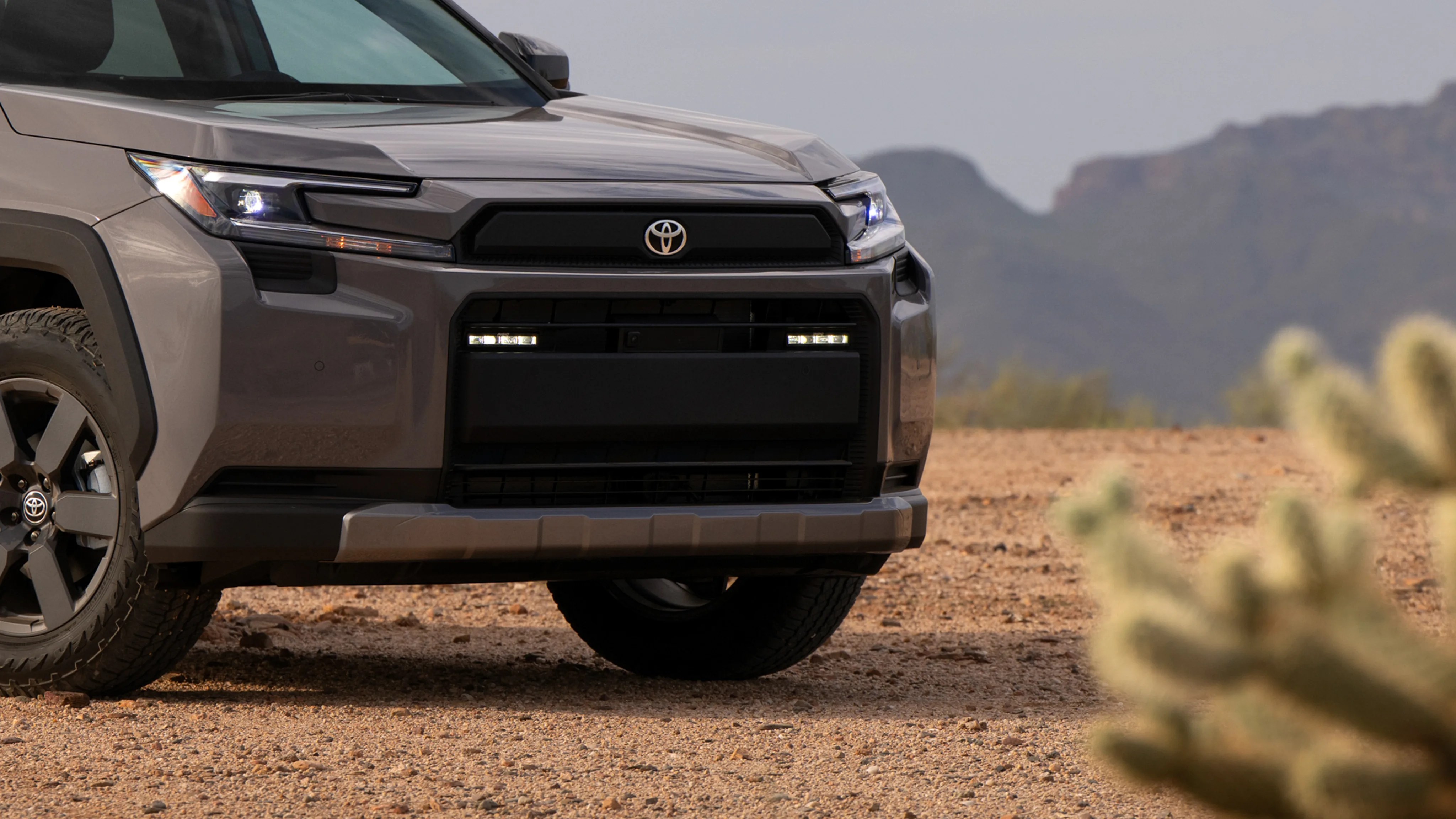 Front view of a gray Toyota SUV with LED headlights on a dirt road with mountains in the background.