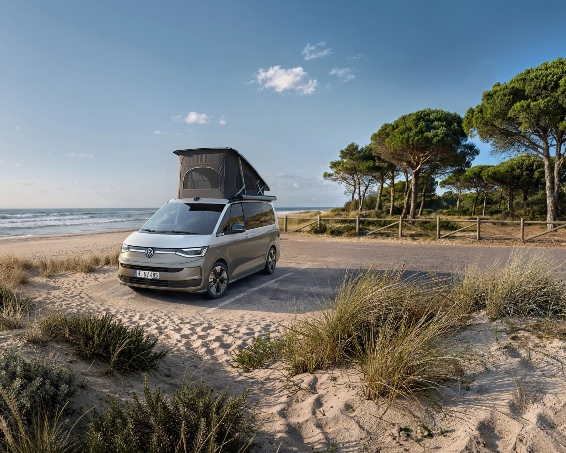 Silver Volkswagen camper van with raised roof tent parked on sandy beach near ocean and pine trees.