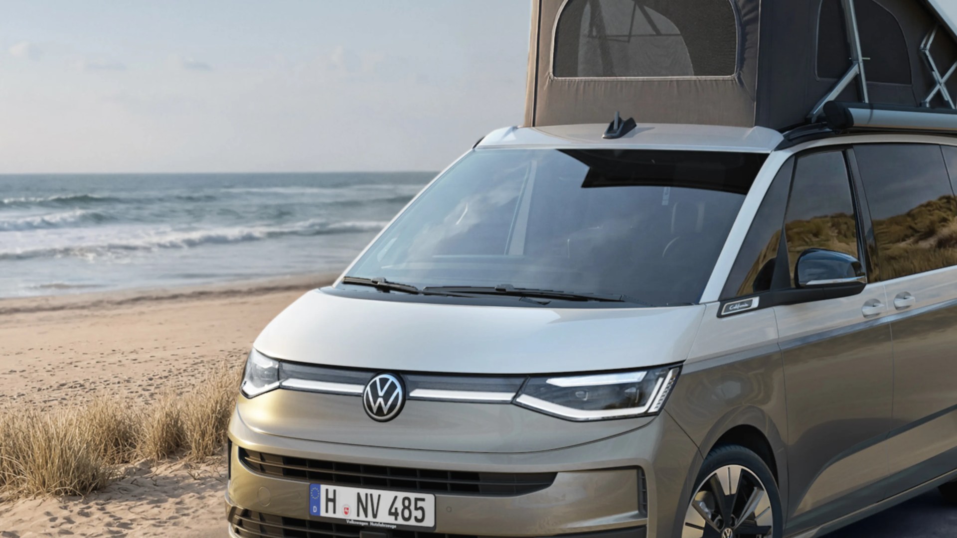 Silver Volkswagen camper van with pop-up roof parked on a sandy beach near the ocean.