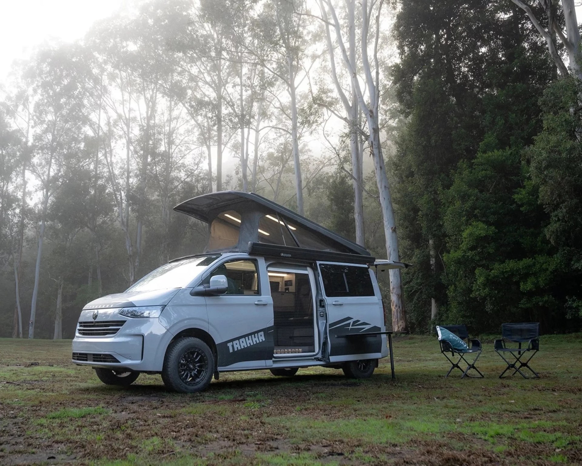 White camper van with pop-up roof and open sliding door parked on grass near trees with two camping chairs nearby.