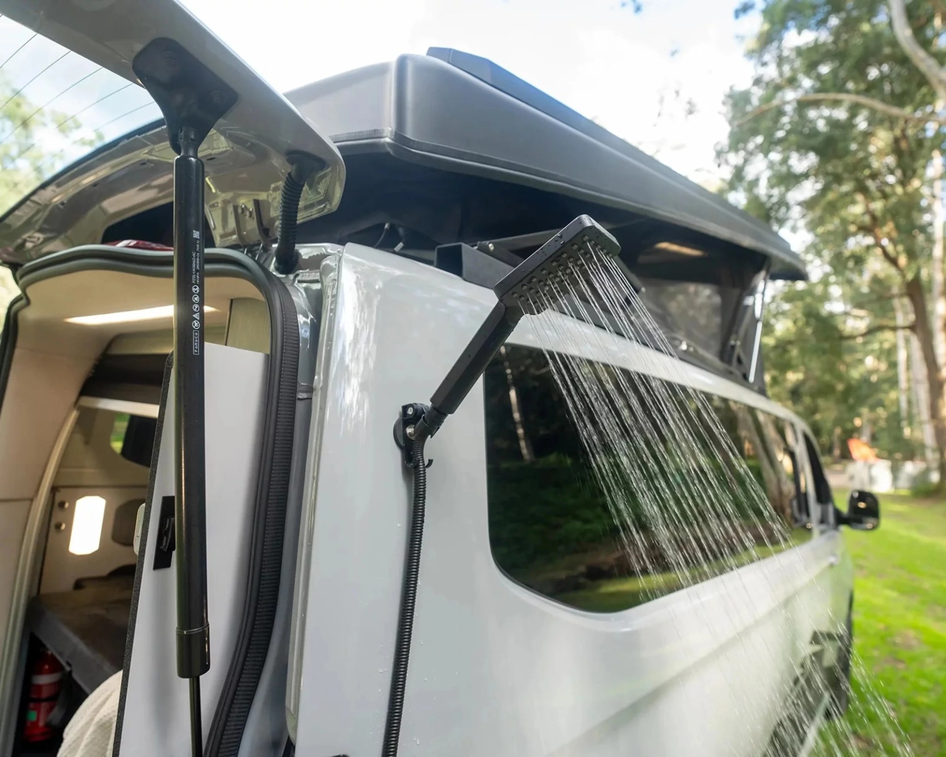 Outdoor shower head spraying water attached to the side of a white camper van with an open door.