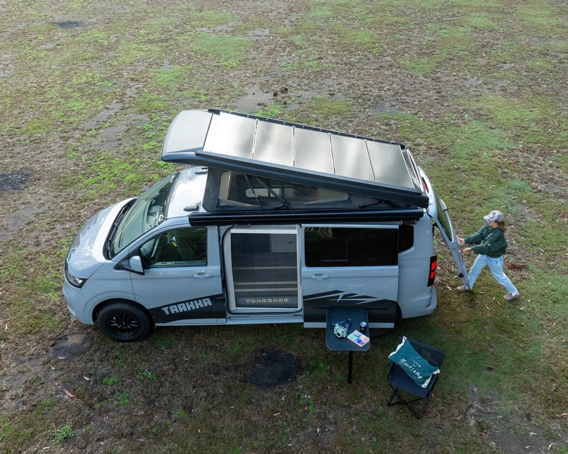 Silver camper van with a raised roof solar panel, open sliding door, small black table with items, and a person near the rear.