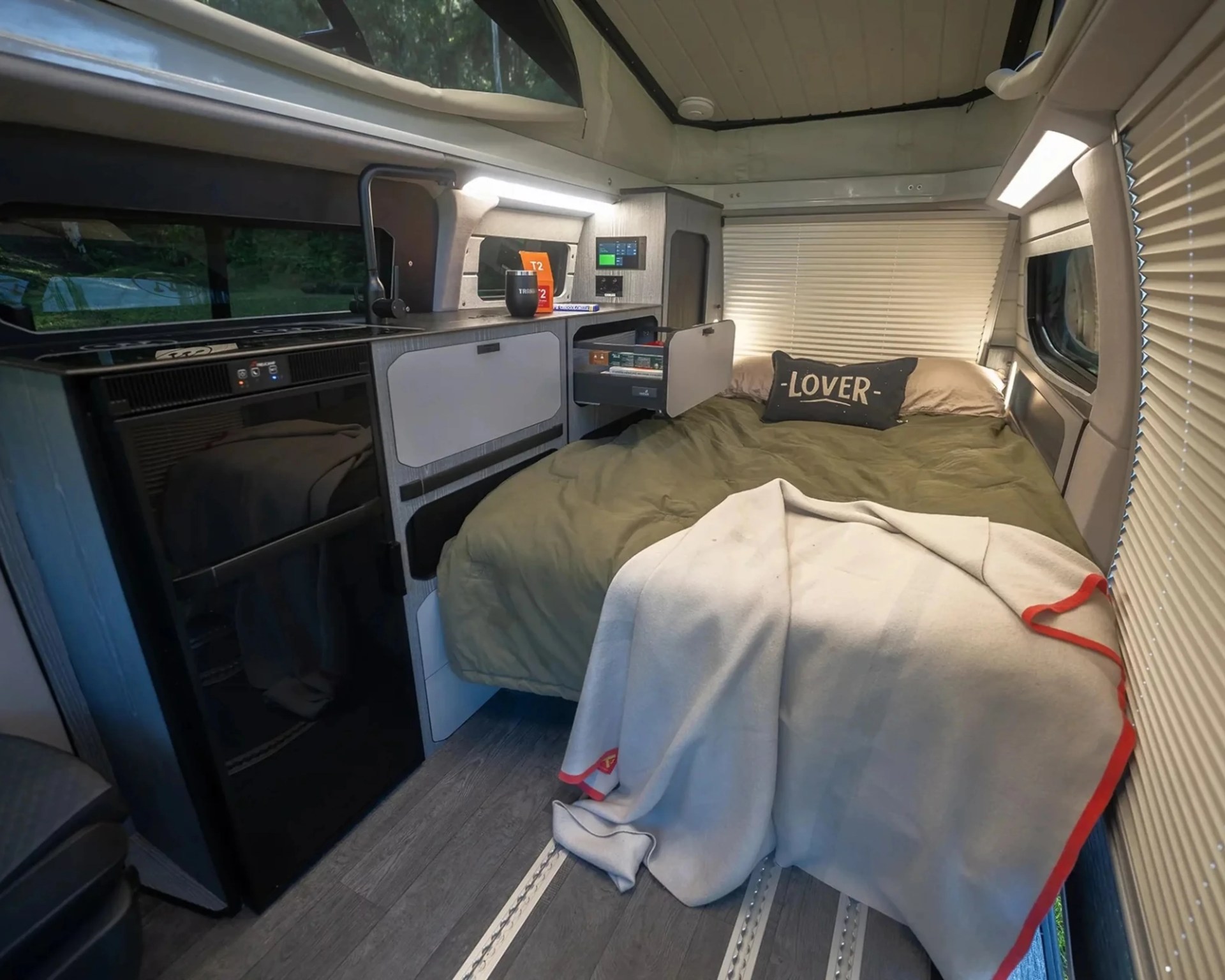 Compact camper van interior with a bed, green bedding, a pillow labeled "LOVER," a small kitchenette, and a gray blanket with red trim.