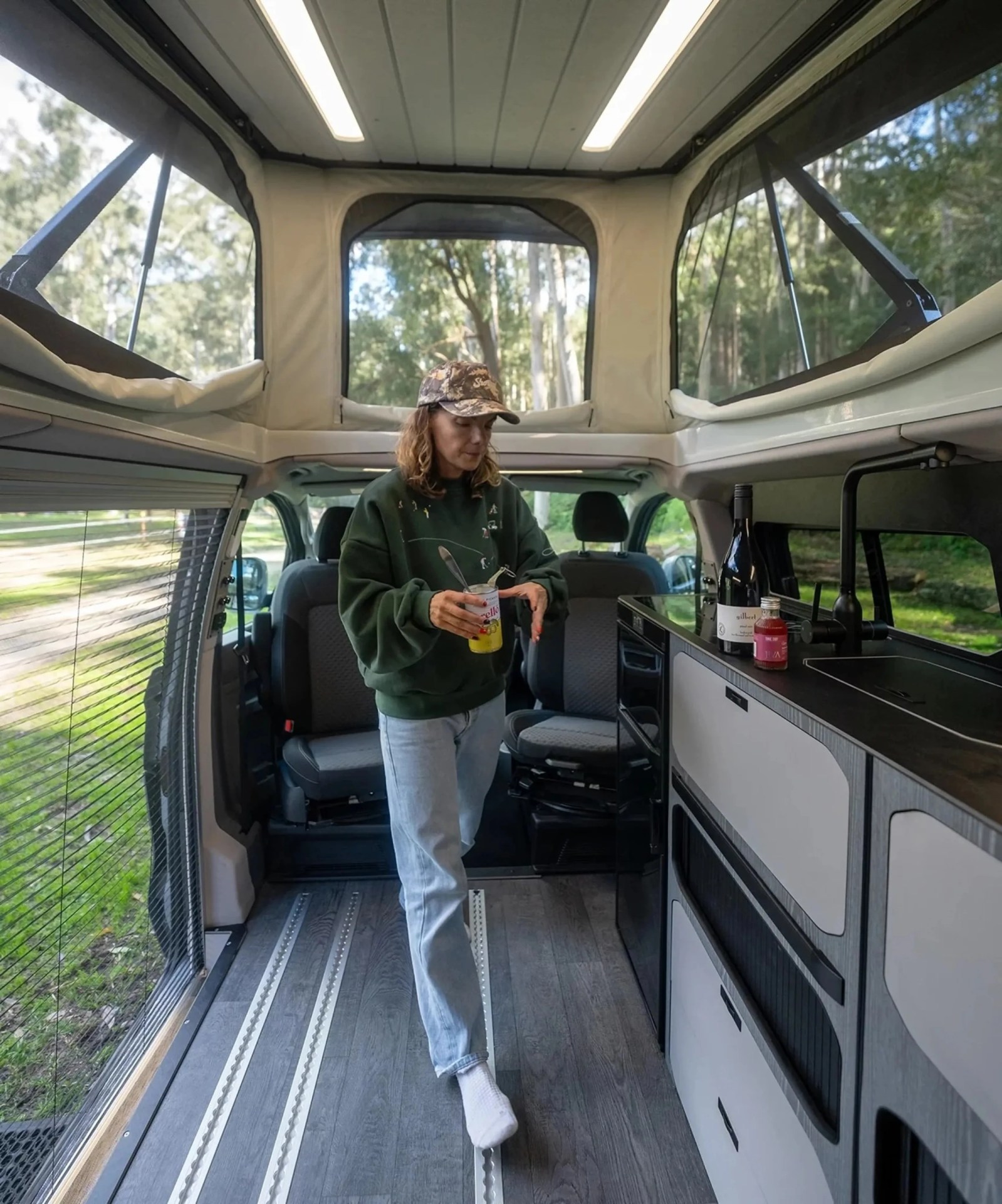 Person in a green sweatshirt and camo cap holding a can inside a camper van with large windows and a kitchenette.